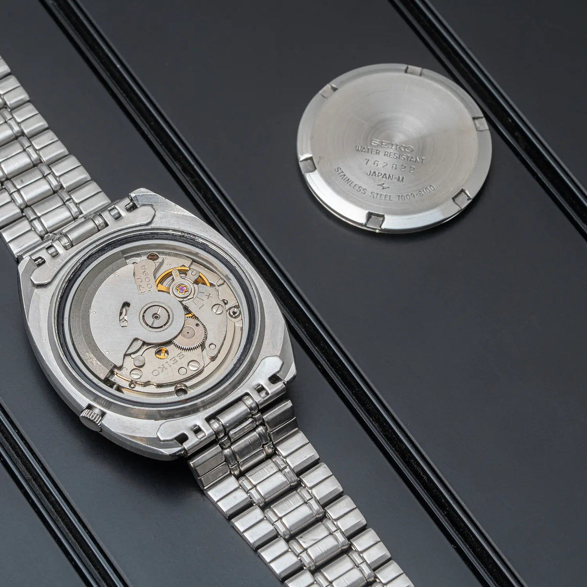 Silver stainless steel watch Seiko, Ref. 7009-8100, Cal. 7009, Men’s Automatic, 1977 with a metal link bracelet, shown with the back case open to reveal the automatic mechanical movement, alongside the detached back cover on a dark textured background.
