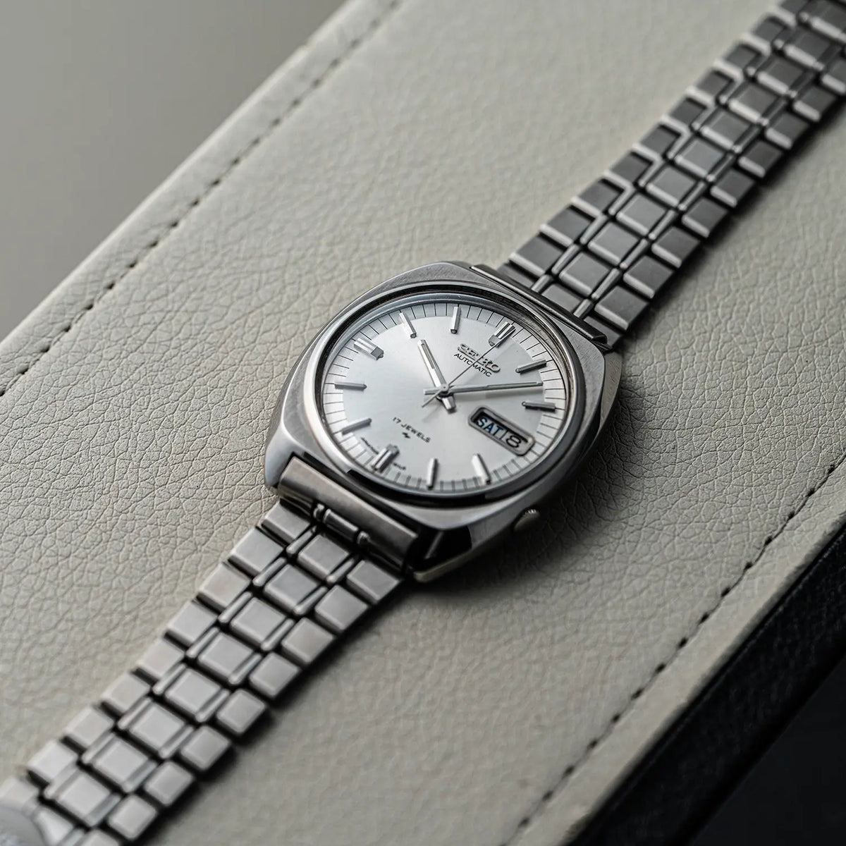 A vintage-style wristwatch Seiko, Ref. 7009-8100, Cal. 7009, Men’s Automatic, 1977 with a silver-toned metal bracelet and a white dial, photographed diagonally on a light gray textured leather surface.