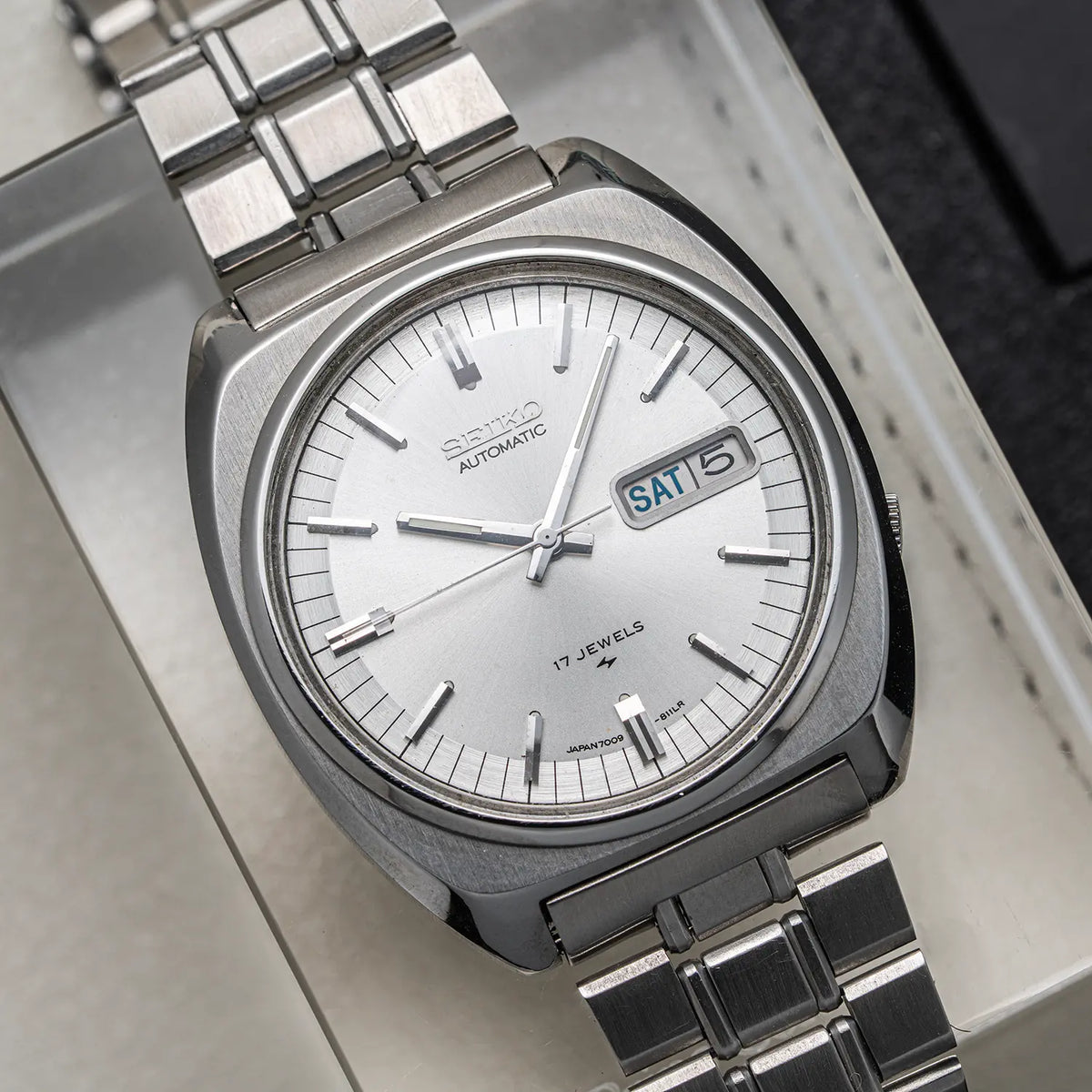 A close-up shot of a vintage Seiko, Ref. 7009-8100, Cal. 7009, Men’s Automatic, 1977 watch with a silver-toned stainless steel case, silver dial, and matching steel link bracelet, resting on a light-colored textured surface.