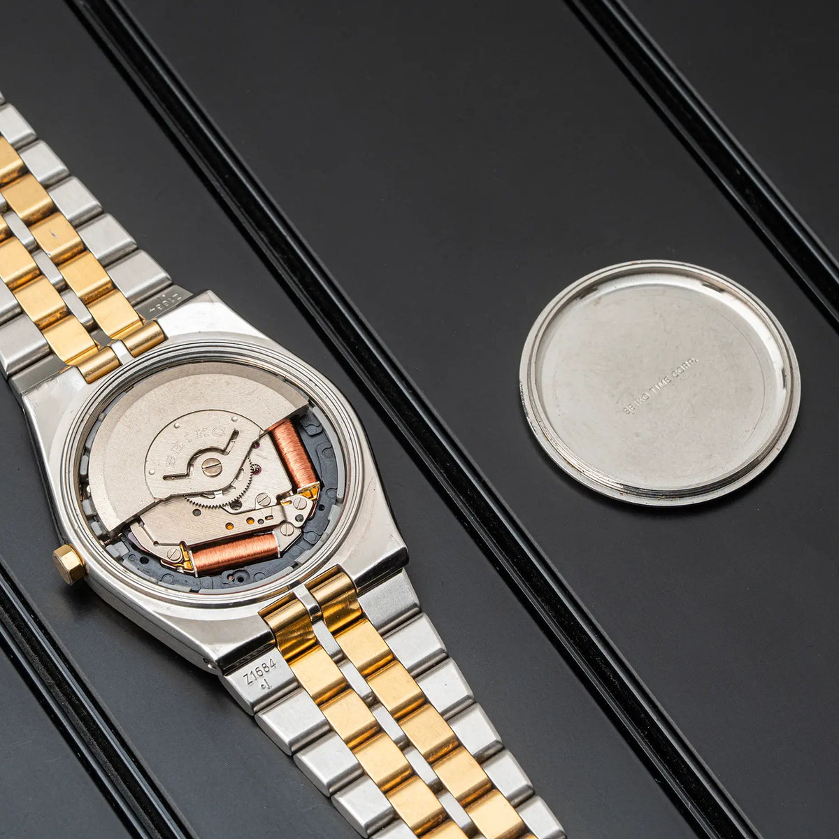 A two-tone stainless steel and gold wristwatch shown open with the case removed, revealing the mechanical movement inside. The matching metal link bracelet is laid diagonally on a dark textured surface, with the detached round case placed beside it.