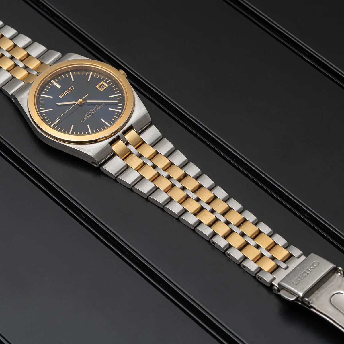 A Seiko AGS, Ref. 5M22-8A10, Cal. 5M22, Men’s Quartz Analog two-tone stainless steel and old wristwatch with a dark blue dial, presented diagonally on a black textured background.