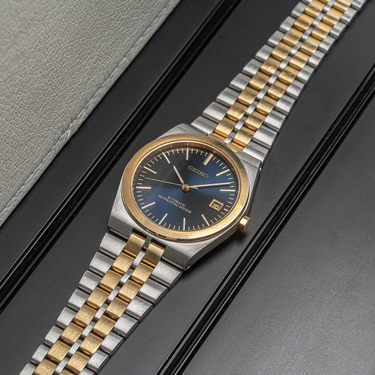 A Seiko AGS, Ref. 5M22-8A10, Cal. 5M22, Men’s Quartz Analog stainless steel and gold-tone two-tone watch with a blue dial, presented diagonally on a dark textured surface next to a light gray textured leather-like surface.