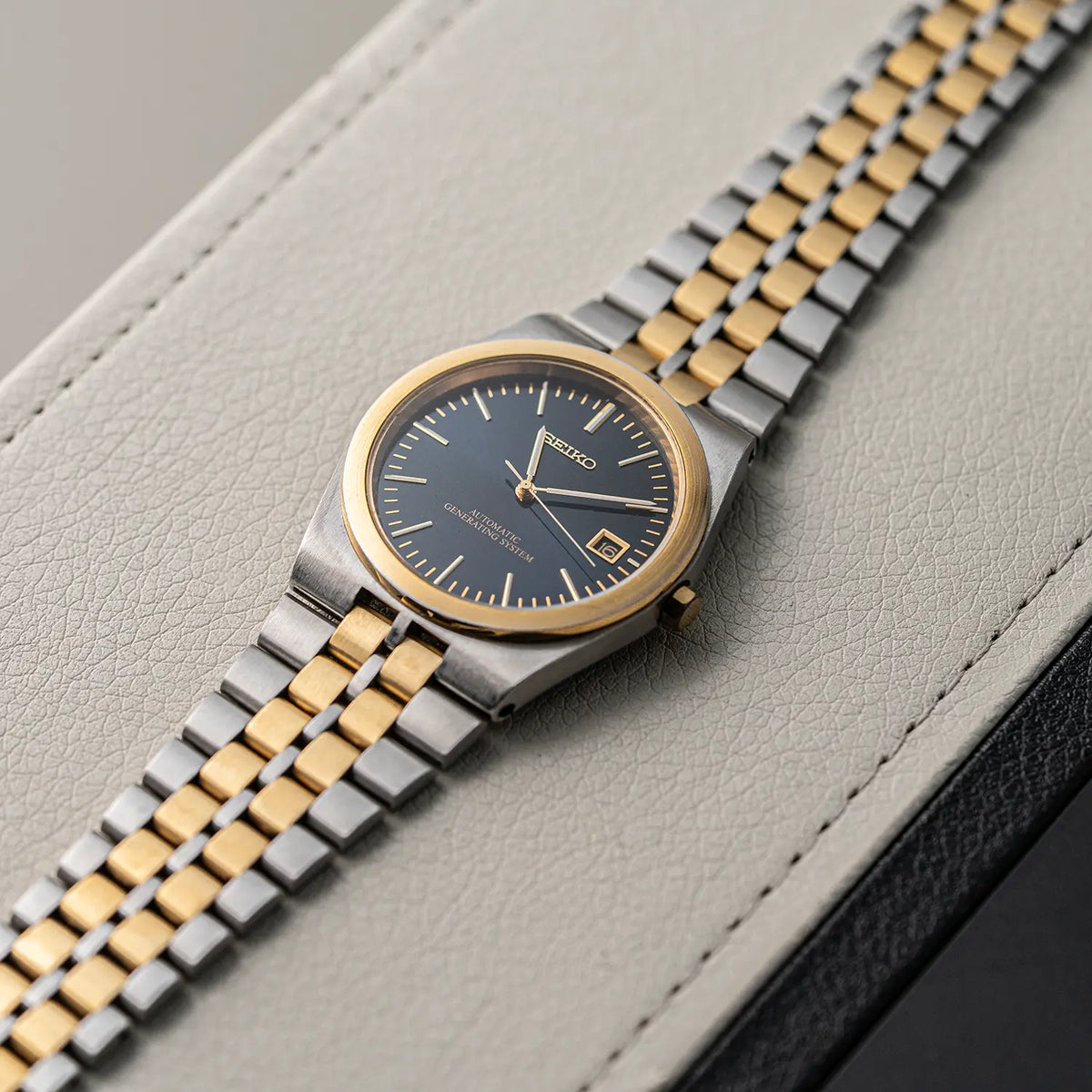 A Seiko AGS, Ref. 5M22-8A10, Cal. 5M22, Men’s Quartz Analog wristwatch with a two-tone silver and gold metal bracelet, a gold-tone case, and a dark blue dial, resting diagonally on a textured light gray leather surface.