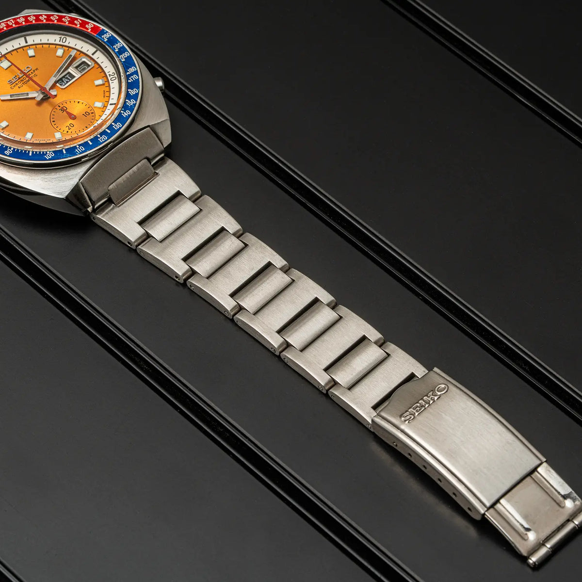 A close-up of a Seiko Chronograph Pogue, Ref. 6139-6002, Cal. 6139, Men’s Chronograph watch with a silver stainless steel link bracelet and a distinctive orange dial, resting on a black textured surface.