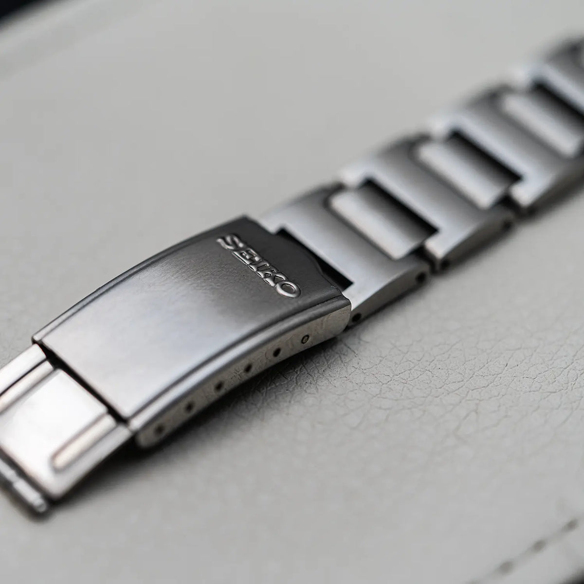 Close-up of a stainless steel watch band clasp with the brand name 'ORAS' engraved on the metal, resting on a textured light gray surface.