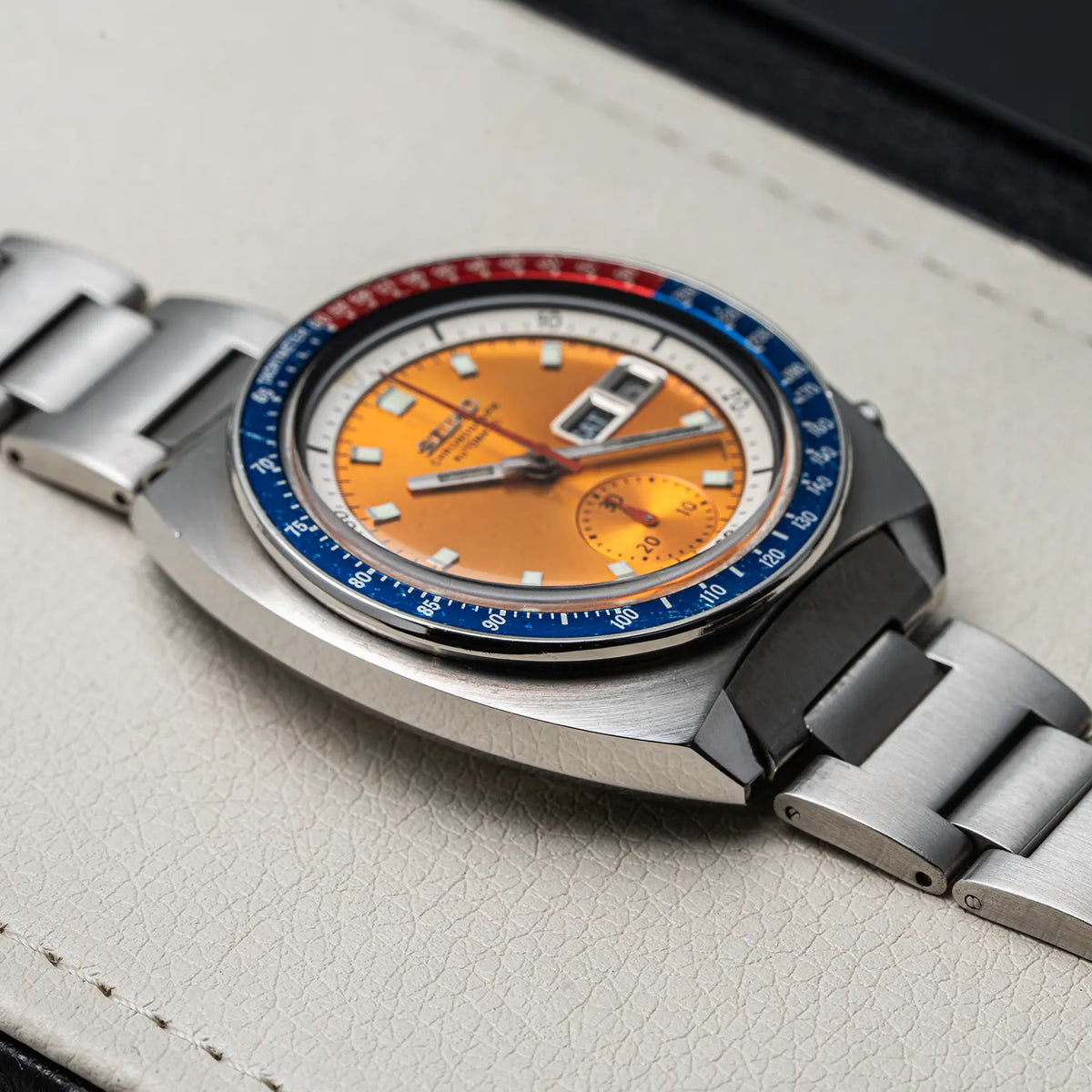 A close-up of a wristwatch Seiko Chronograph Pogue, Ref. 6139-6002, Cal. 6139, Men’s Chronograph with a brushed silver-tone metal bracelet and case, featuring a vibrant orange dial and a blue-and-red bezel, resting on a textured light beige leather surface.
