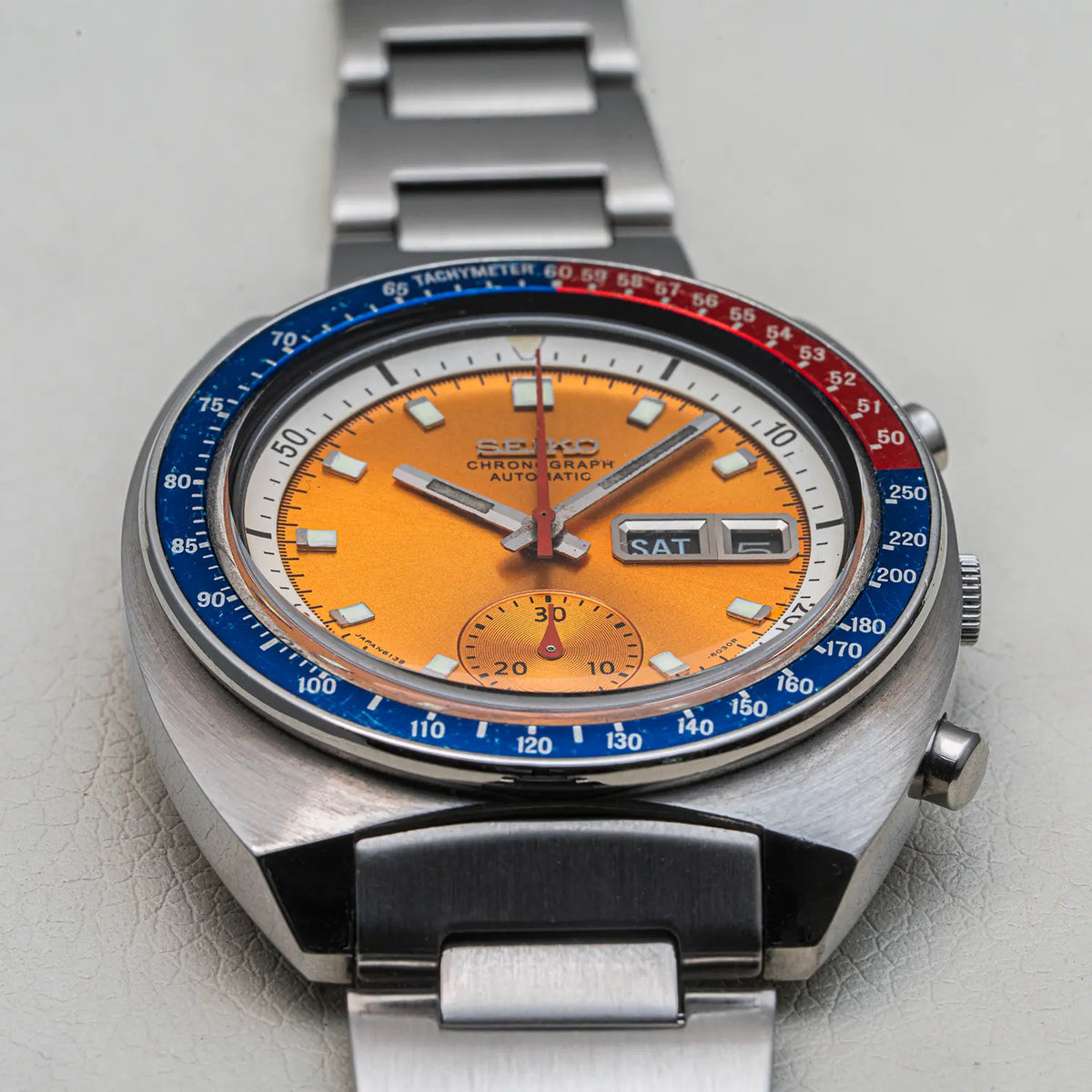 A Seiko Chronograph Pogue, Ref. 6139-6002, Cal. 6139, Men’s Chronograph watch with a distinctive orange dial, a blue and red tachymeter bezel, and a stainless steel link bracelet, shown on a light textured background.
