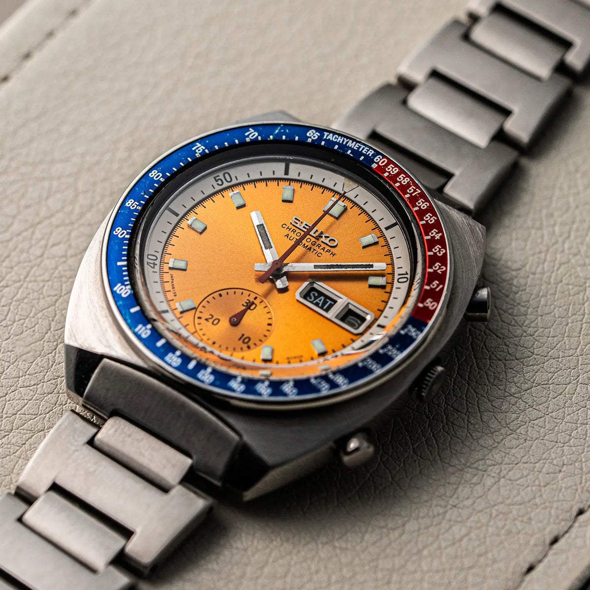 A close-up shot of a Seiko Chronograph Pogue, Ref. 6139-6002, Cal. 6139, Men’s Chronograph wristwatch with a distinctive orange dial, blue and red bezel, and a stainless steel link bracelet, resting on a textured gray leather surface.