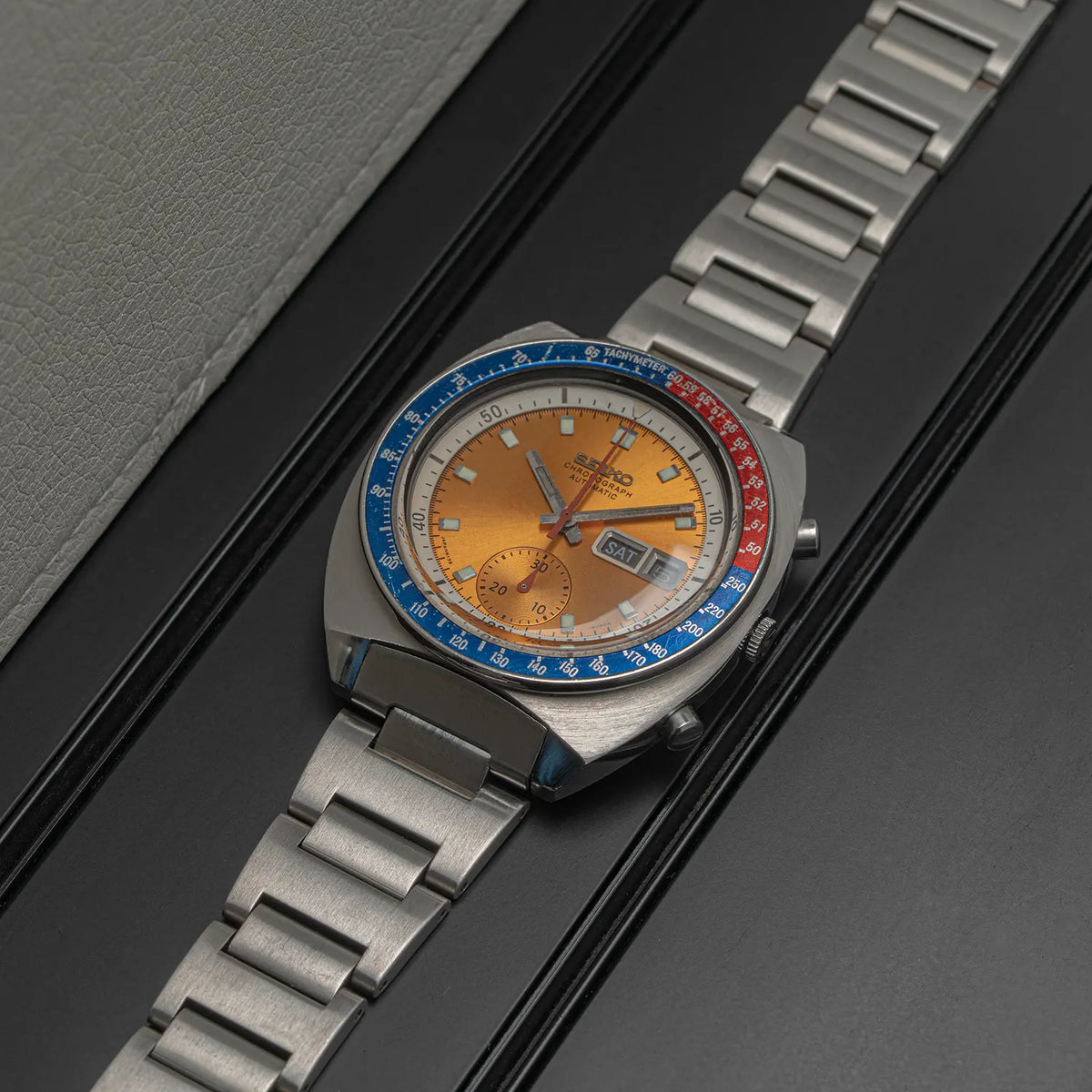 A vintage-style wristwatch Seiko Chronograph Pogue, Ref. 6139-6002, Cal. 6139, Men’s Chronograph with a stainless steel link bracelet, a brushed silver-tone case, and a distinctive orange dial. The bezel features a split blue and red racing-style track layout.