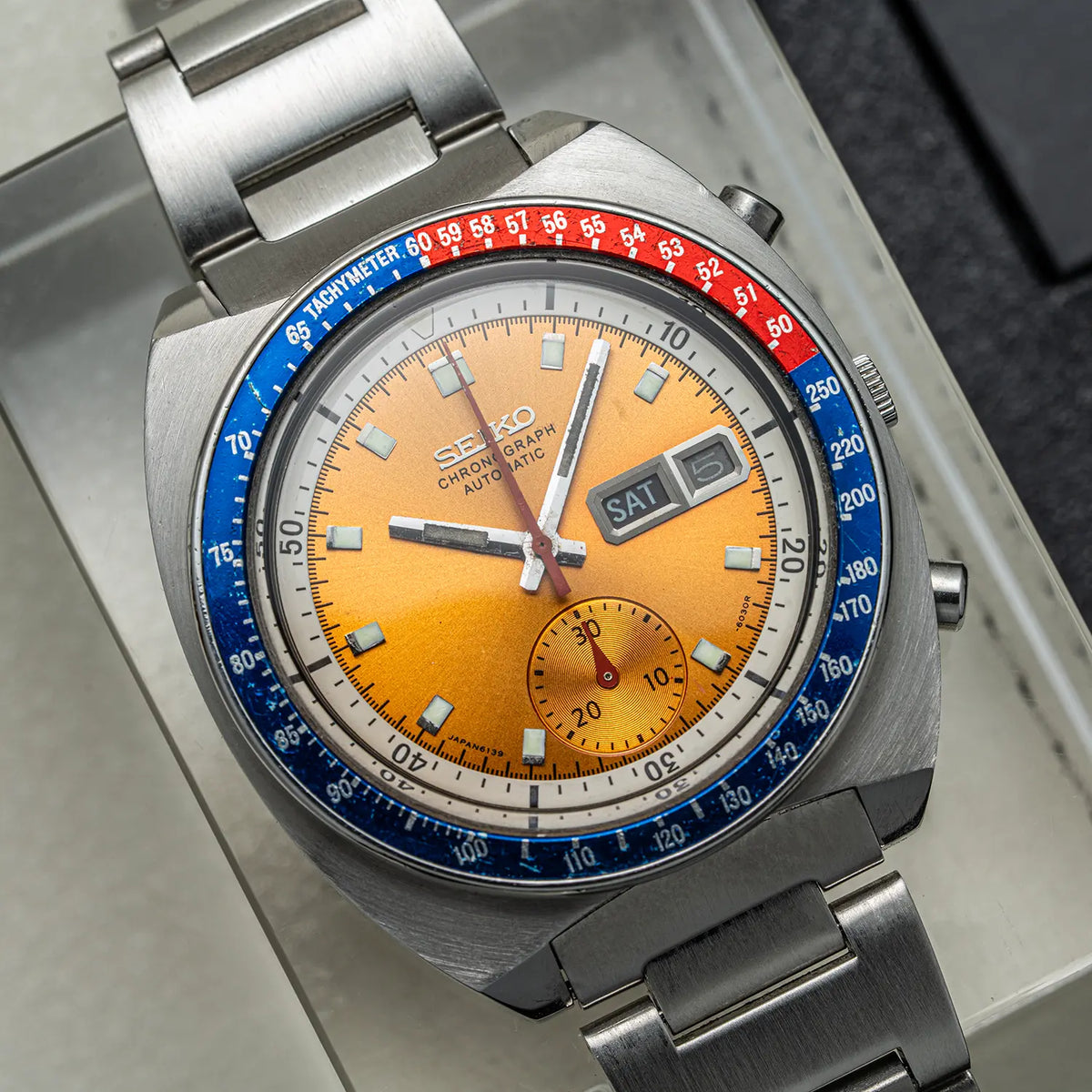 A close-up shot of a Seiko Chronograph Pogue, Ref. 6139-6002, Cal. 6139, Men’s Chronograph Automatic watch with a distinctive orange dial, a blue and red tachymeter bezel, and a stainless steel link bracelet.