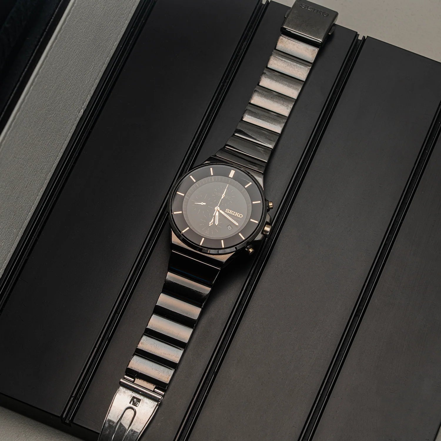 A Seiko Chronograph, Ref. 7T92-0NGO stainless steel watch with a dark dial and matching dark metal link bracelet, presented diagonally on a black textured box.