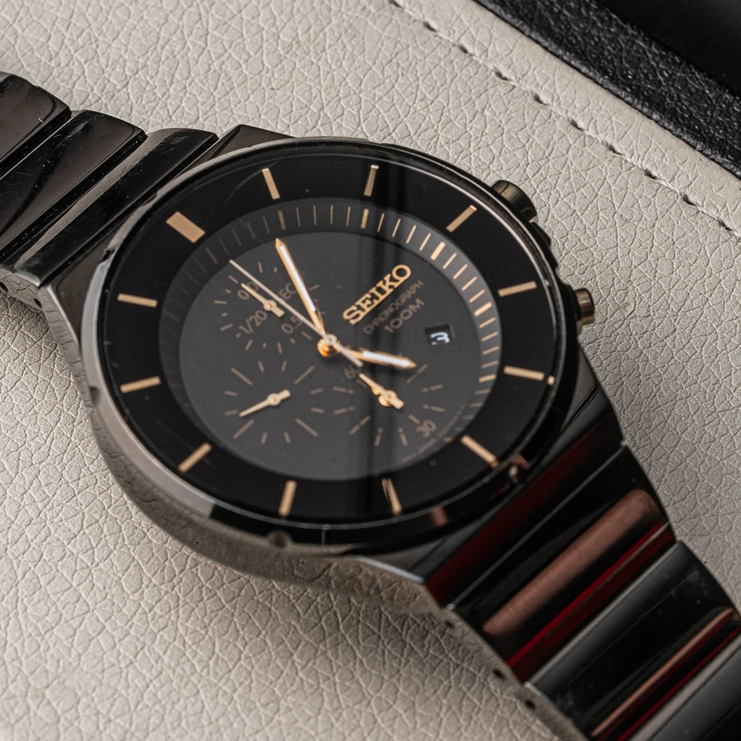 A close-up of a Seiko Chronograph, Ref. 7T92-0NGO analog watch with a black dial, black metal case, and a dark metallic link bracelet with a reddish-brown sheen, resting on a textured light gray surface.