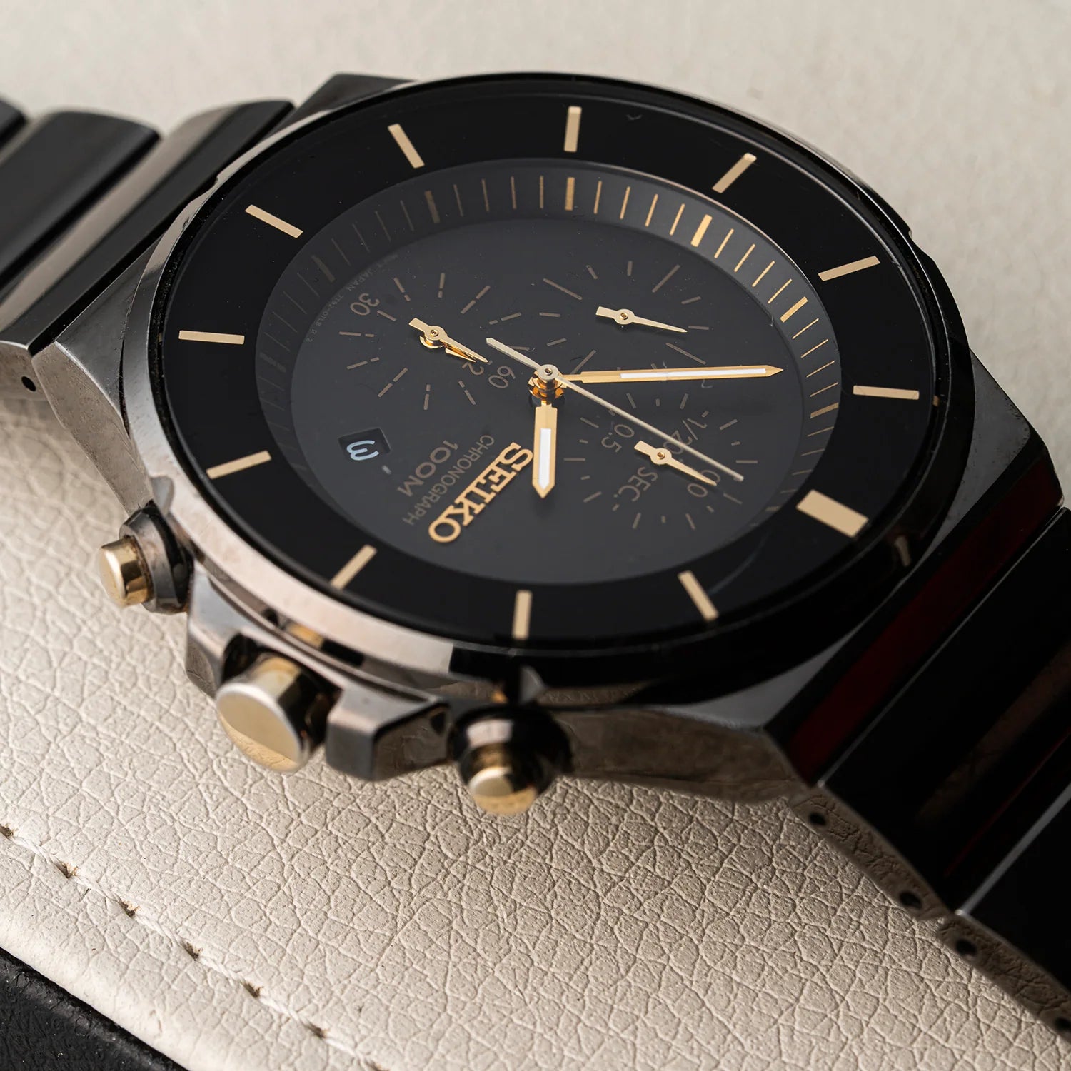 A close-up of a Seiko Chronograph, Ref. 7T92-0NGO analog chronograph watch with a black dial, black bezel, and a dark metallic case accented by gold-tone details and pushers, resting on a textured light beige leather surface.