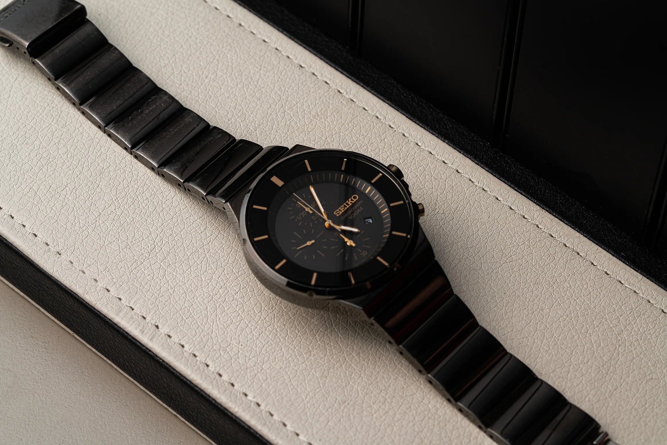 A wristwatch Seiko Chronograph, Ref. 7T92-0NGO with a black ceramic link bracelet and a black dial featuring gold-tone hands and markers, resting diagonally on a textured off-white leather surface.