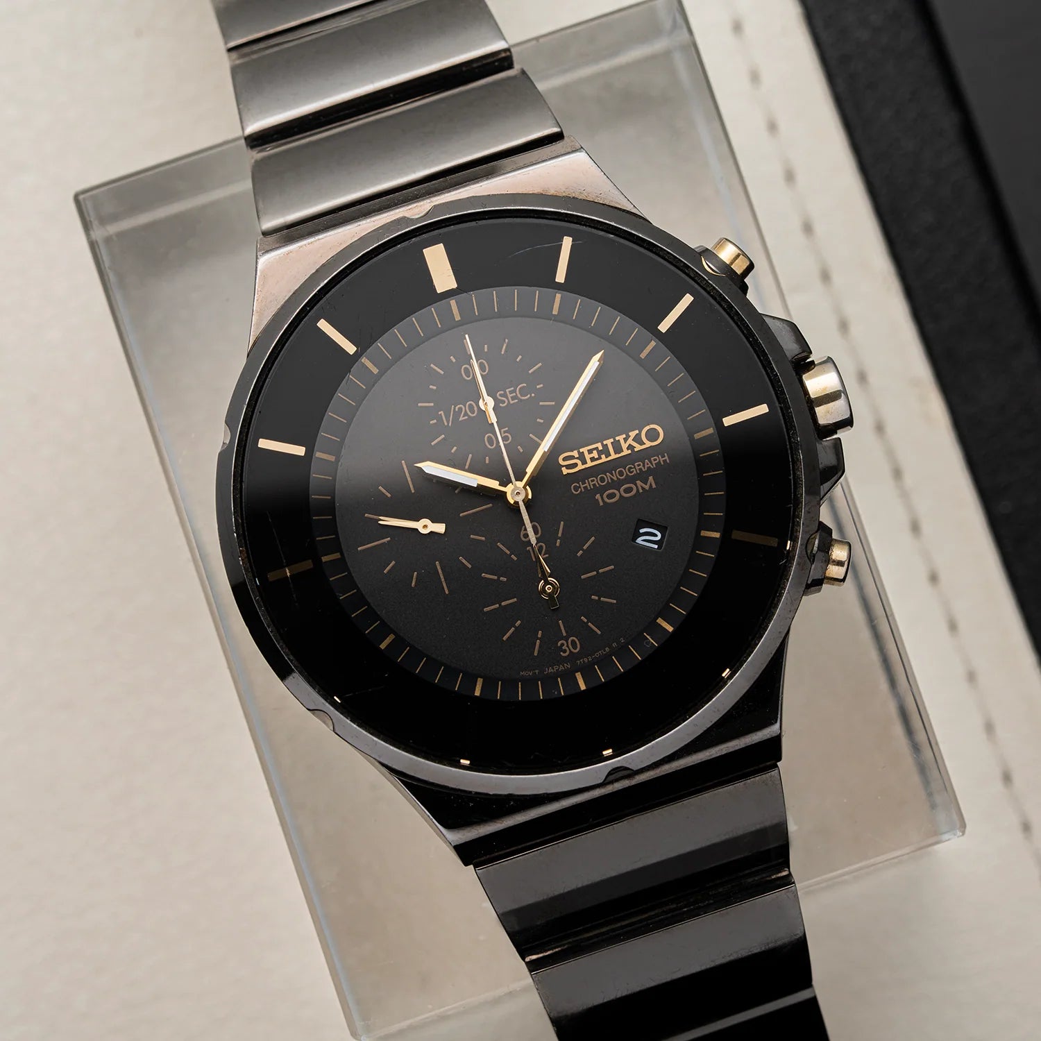 A Seiko Chronograph, Ref. 7T92-0NGO watch with a black dial, black and silver-tone metal case, and black metal link bracelet, displayed on a clear acrylic stand against a light neutral background.