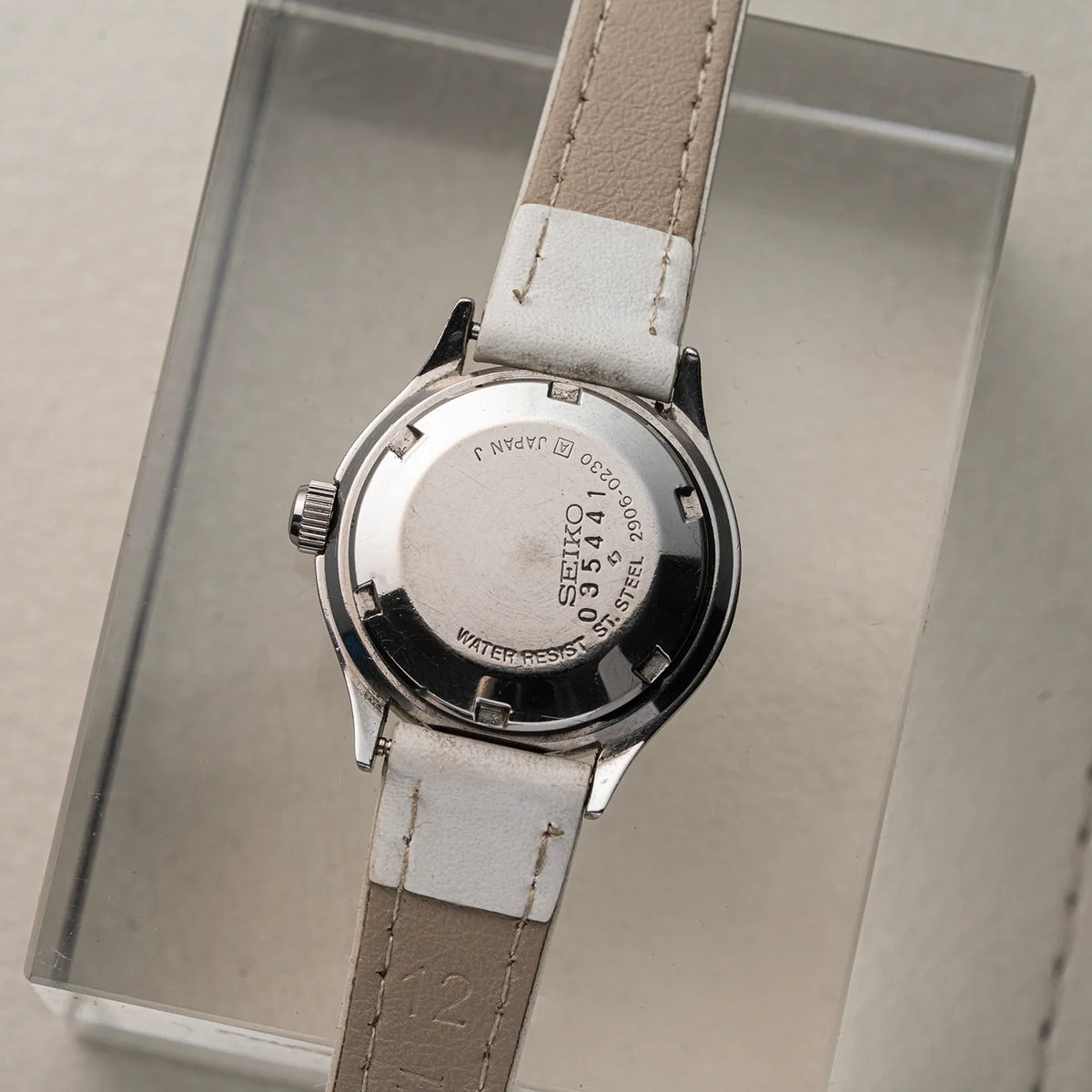 A vintage Seiko, Ref. 2906-0230 women's watch with a silver-toned metal case and a light beige leather strap, shown from the back resting on a clear acrylic display block.