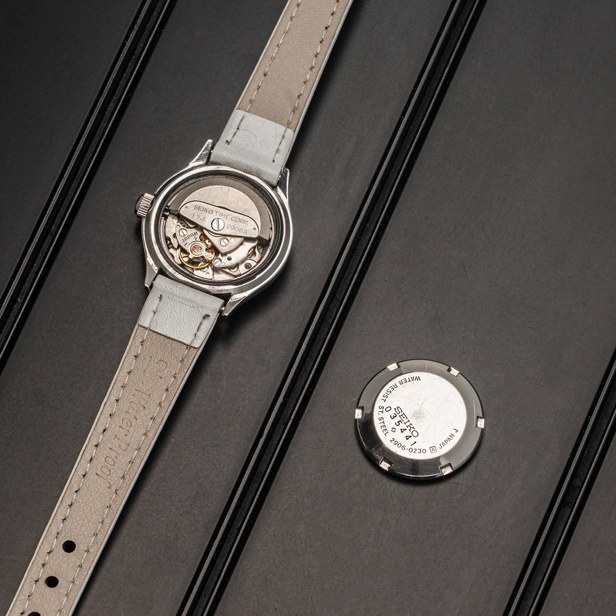 A silver-tone automatic watch Seiko, Ref. 2906-0230 with a light beige leather strap, shown with its case back open and a separate circular backplate placed beside it on a dark textured surface.