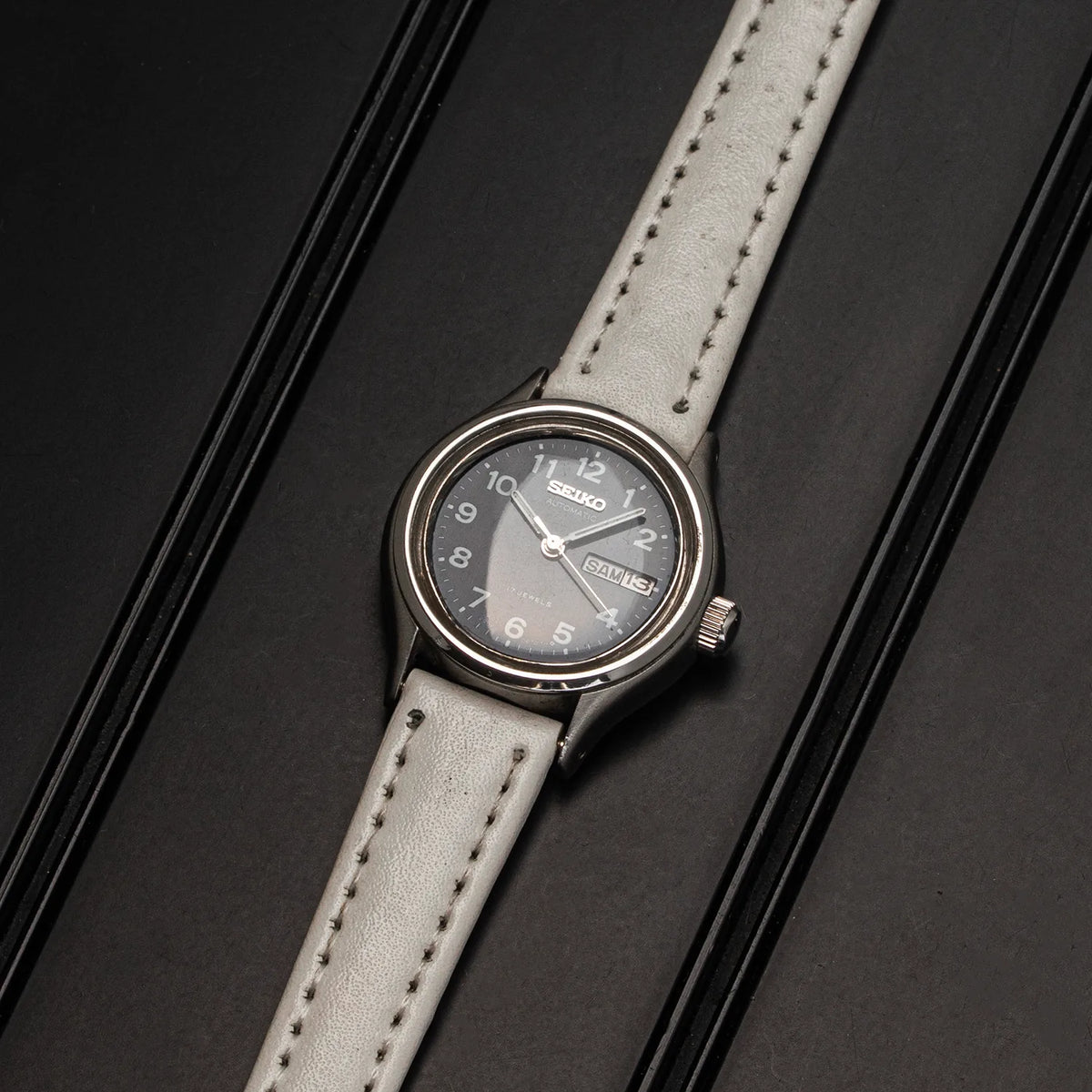 A Seiko, Ref. 2906-0230 women's watch  with a silver-toned case, a dark gray dial, and a light gray leather strap, presented diagonally on a black textured background.