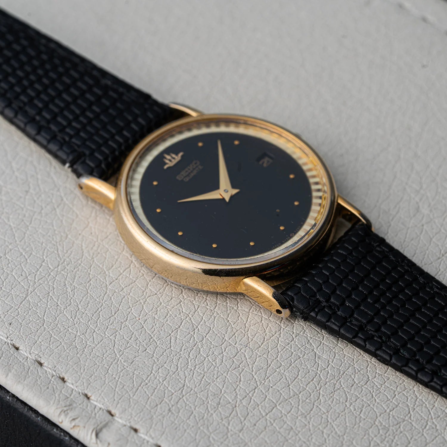 A vintage wristwatch Seiko, Ref. 5Y39-7010 with a gold-tone case, black dial, and black textured strap, resting on a light gray textured surface.