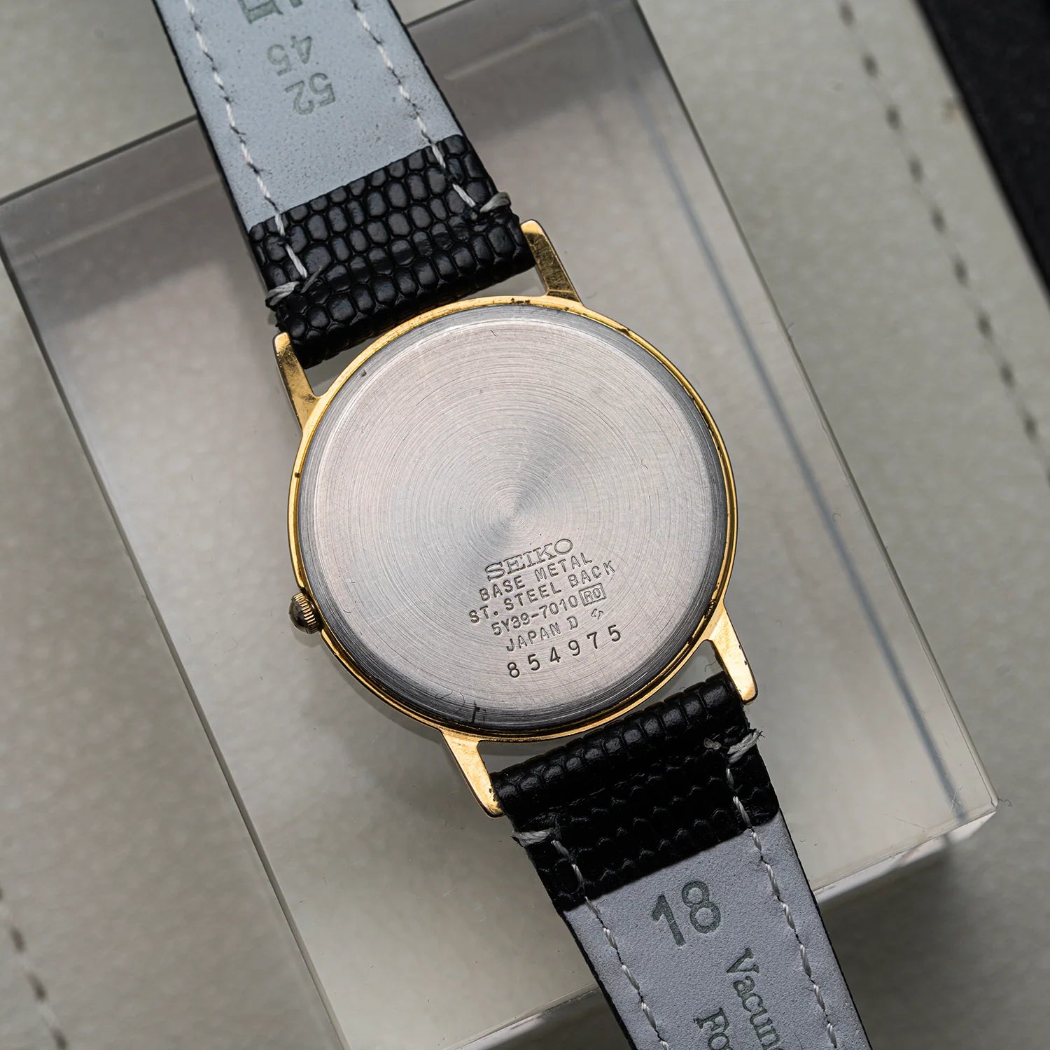 A vintage Seiko watch shown from the back, featuring a brushed stainless steel case, gold-tone lugs, and a black textured leather strap on a light gray presentation box.