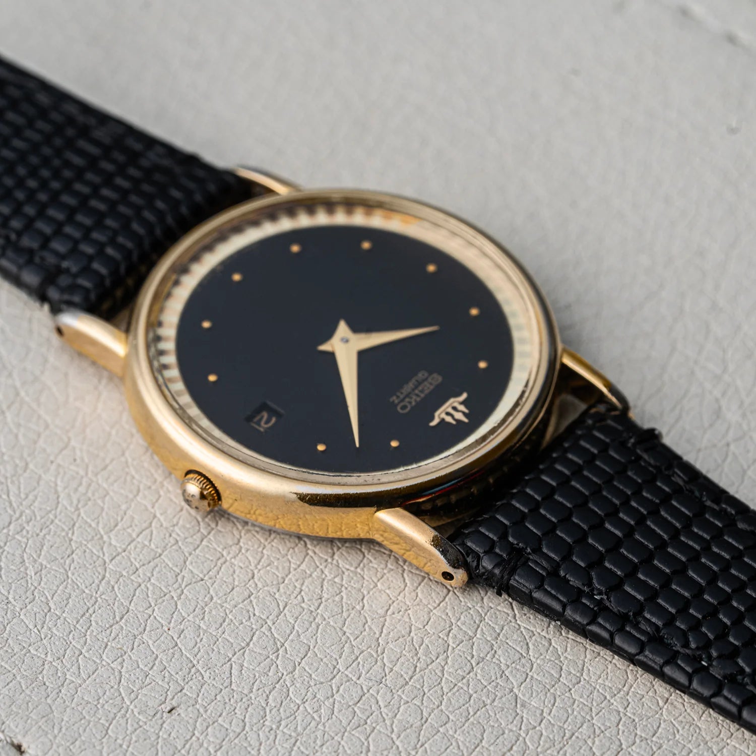 A vintage Seiko, Ref. 5Y39-7010 watch with a gold-tone case, black dial, and black textured leather strap, photographed diagonally on a light textured surface.