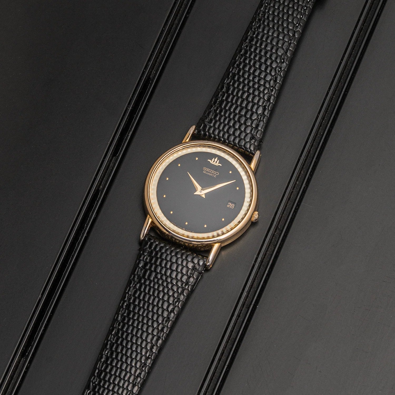 A vintage-style wristwatch Seiko, Ref. 5Y39-7010 with a round gold-tone case, black dial, and textured black strap, photographed diagonally on a dark glossy surface.