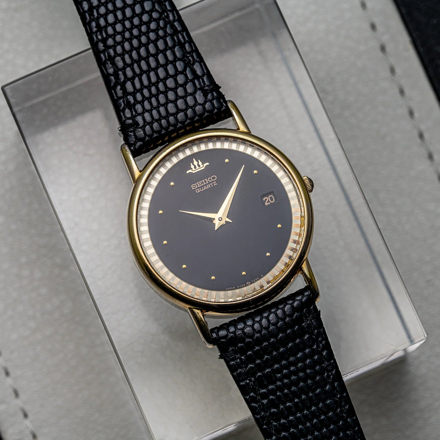 A vintage-style Seiko, Ref. 5Y39-7010 wristwatch with a round gold-tone case, dark charcoal gray dial, and textured black strap, displayed diagonally on a clear acrylic block over a gray surface.