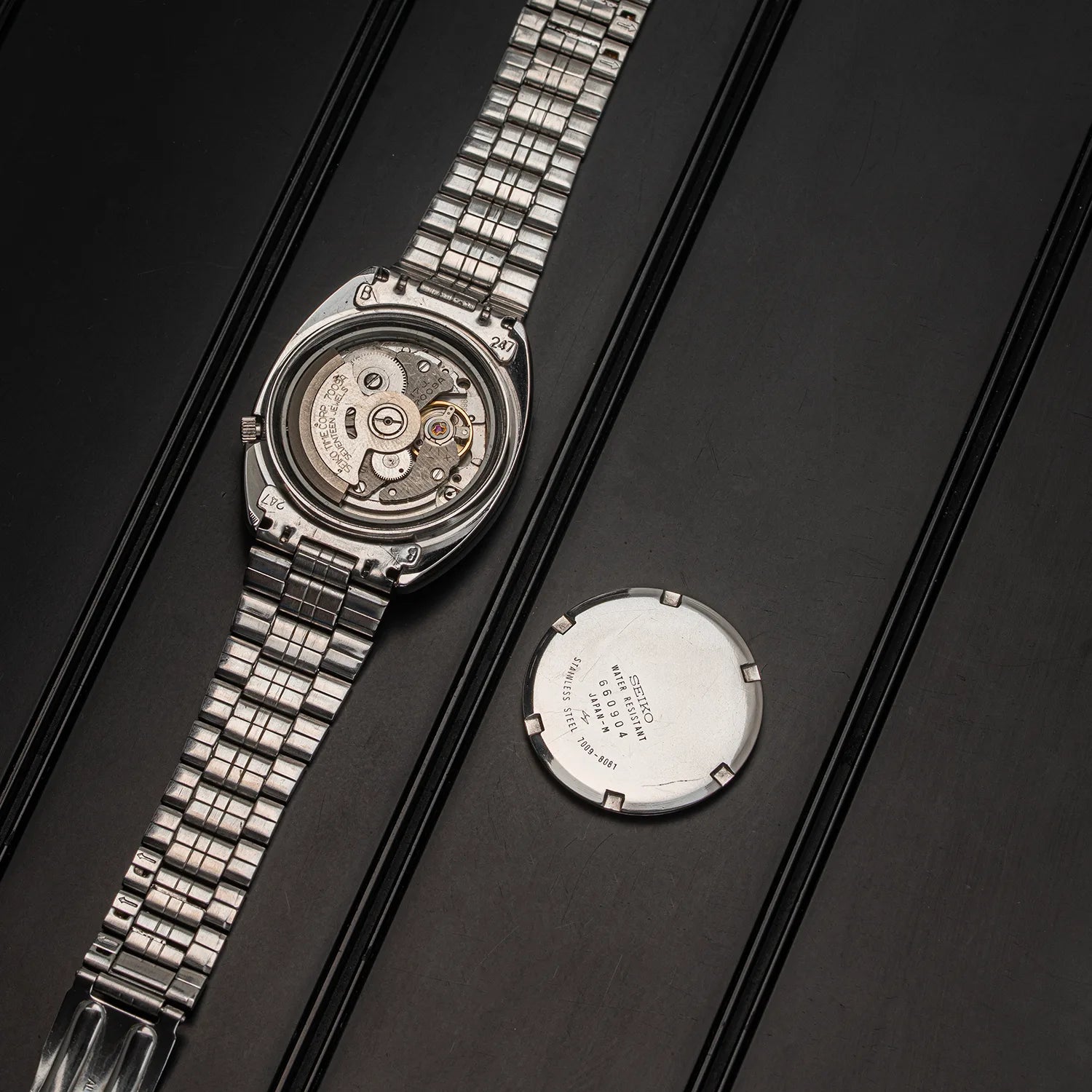 A silver stainless steel wristwatch Seiko 7009-8081 with a transparent case back showing the mechanical movement, displayed alongside its detached round silver backplate on a dark textured background.