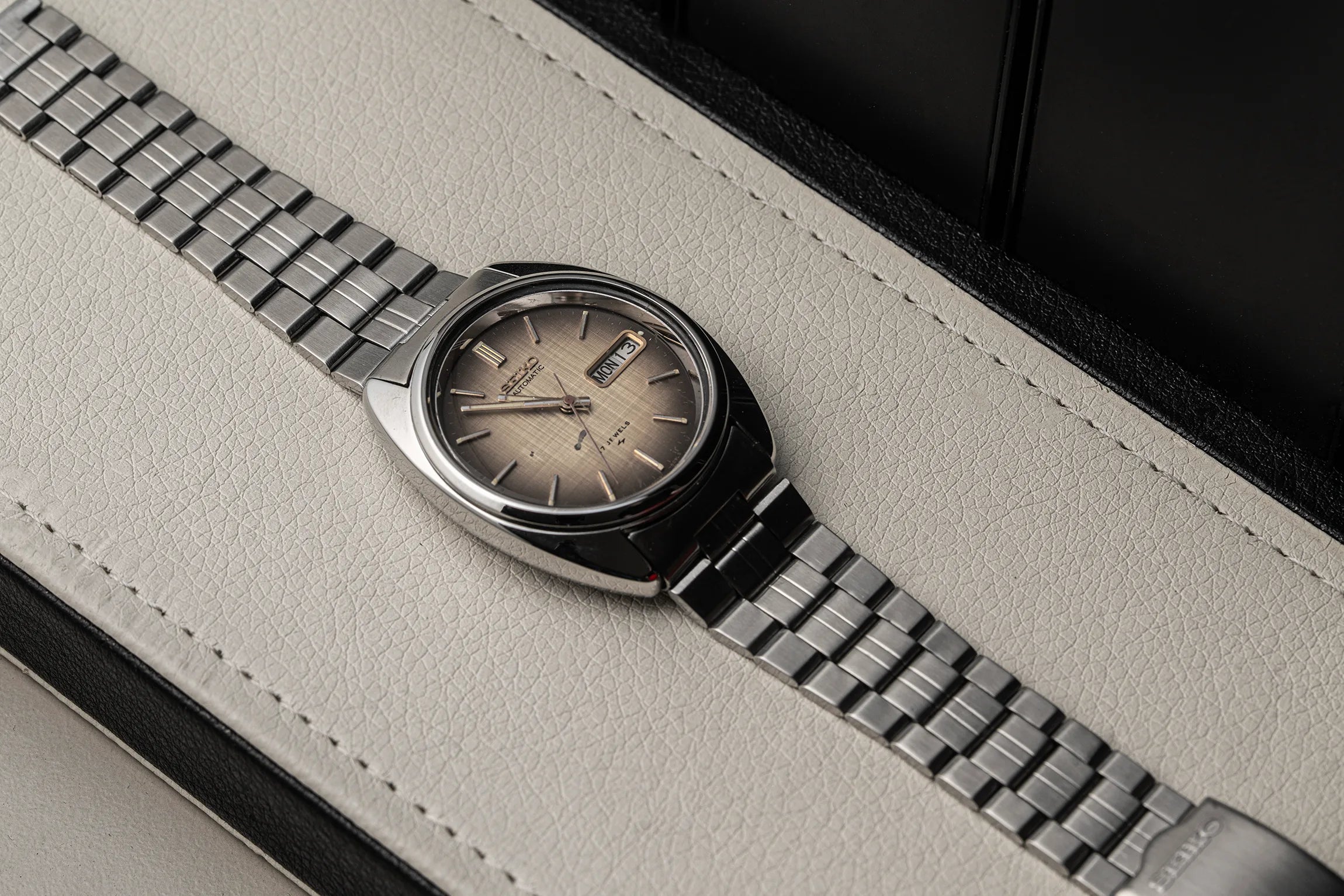 A close-up of a wristwatch Seiko 7009-8081 with a silver-toned metal link bracelet and a champagne-gold dial, resting on a light beige textured leather surface.