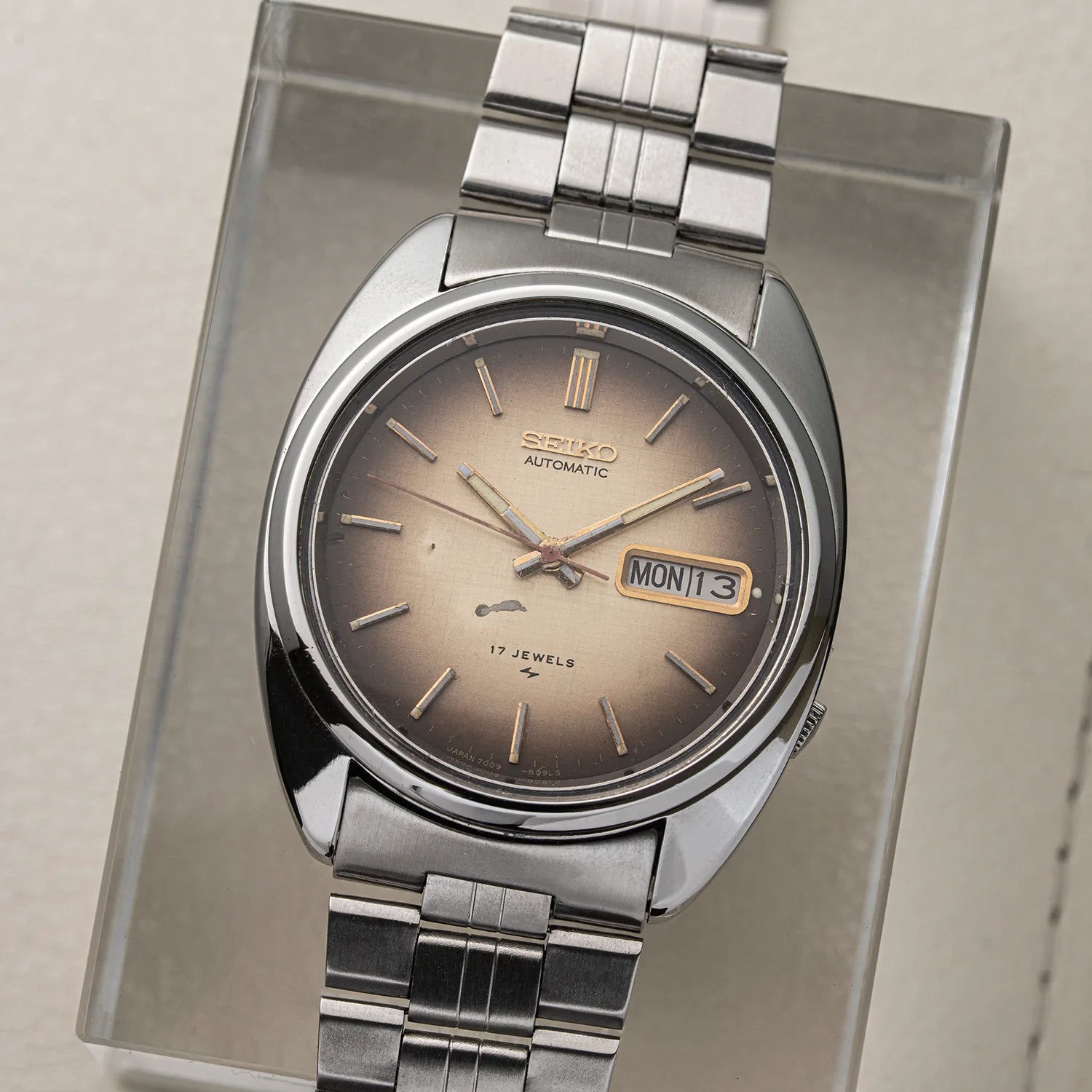A vintage Seiko 7009-8081 automatic watch with a silver-tone metal bracelet and a bronze-brown sunburst dial, displayed on a clear acrylic stand.