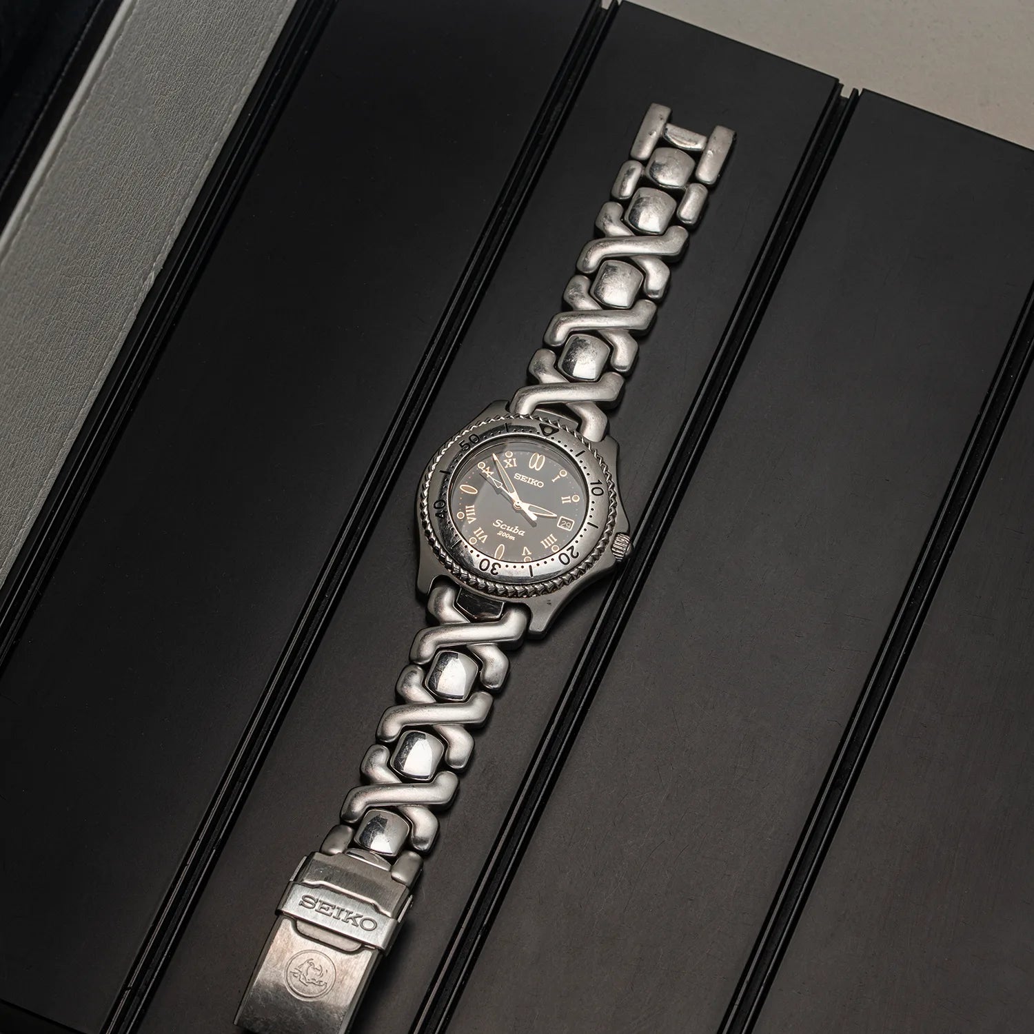 A silver-tone stainless steel watch Seiko Scuba with a textured bezel and matching link bracelet, displayed diagonally on a black presentation box. The brand name 'SEIPRO' is visible on the clasp.
