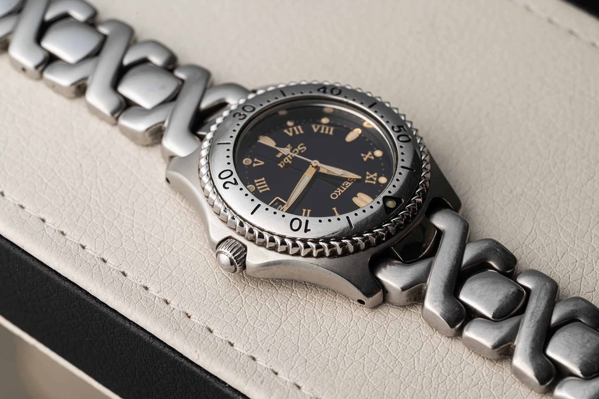 A close-up shot of a Seiko Scuba wristwatch featuring a silver-toned stainless steel link bracelet, a black dial with gold-tone Roman numerals and hands, and a textured bezel, resting on a light beige textured leather surface.