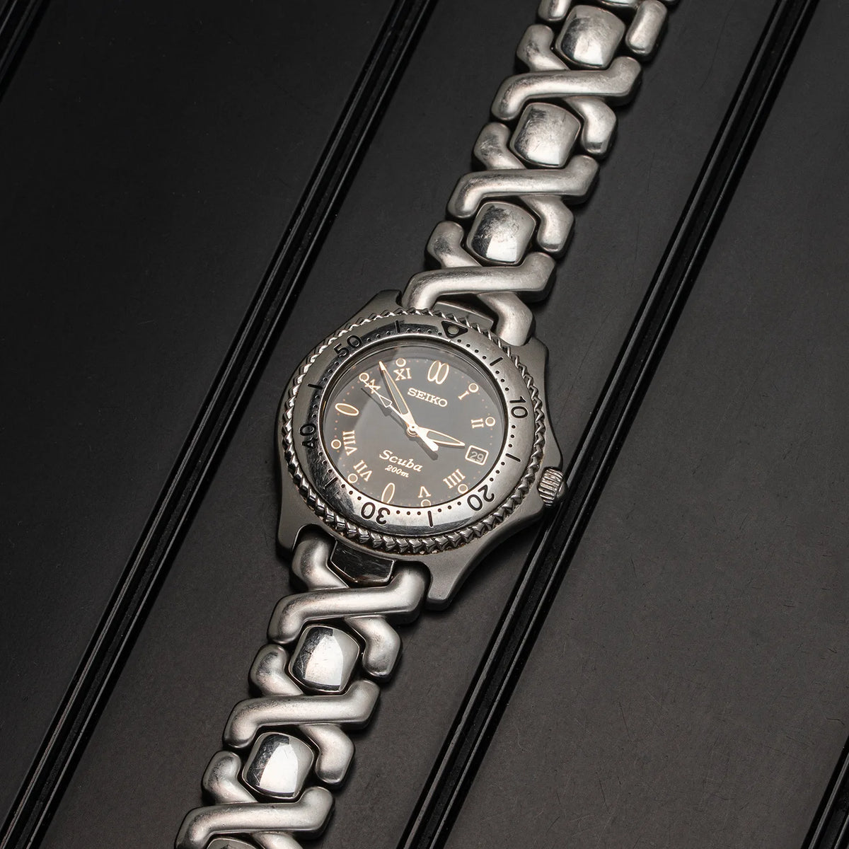 A Seiko Scuba stainless steel watch with a dark gray dial and a distinctive interlocking link bracelet, photographed diagonally on a dark textured background.