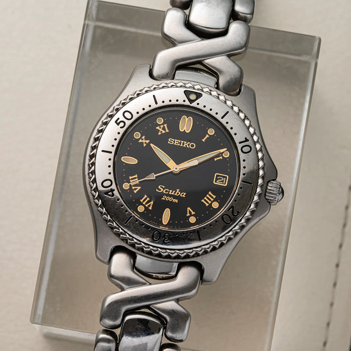 A Seiko Scuba watch with a silver-toned metal link bracelet and a black dial, displayed on a clear acrylic stand.