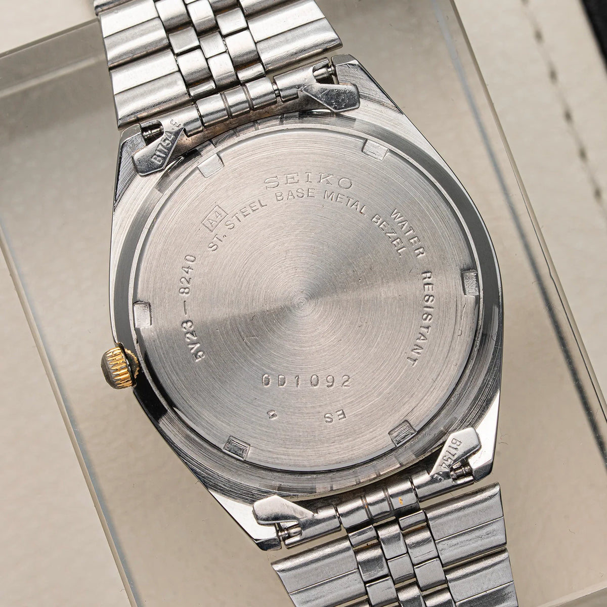 Close-up of the back of a silver stainless steel wristwatch Seiko SQ 5Y23-8240 with a metal link bracelet, resting on a light beige surface. The watch case and bracelet are polished silver with a small gold crown accent visible on the left side.