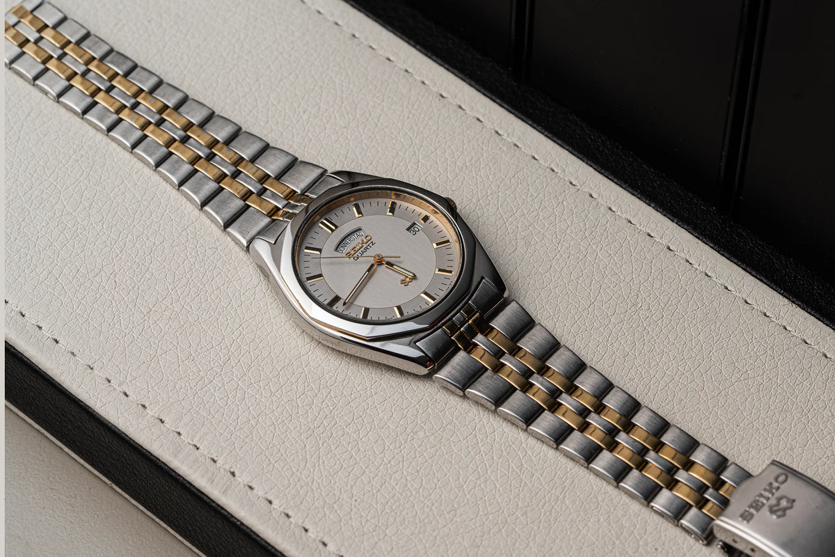 A two-tone stainless steel and gold watch Seiko SQ 5Y23-8240 with a silver dial, resting diagonally on a textured off-white leather surface with a black background.