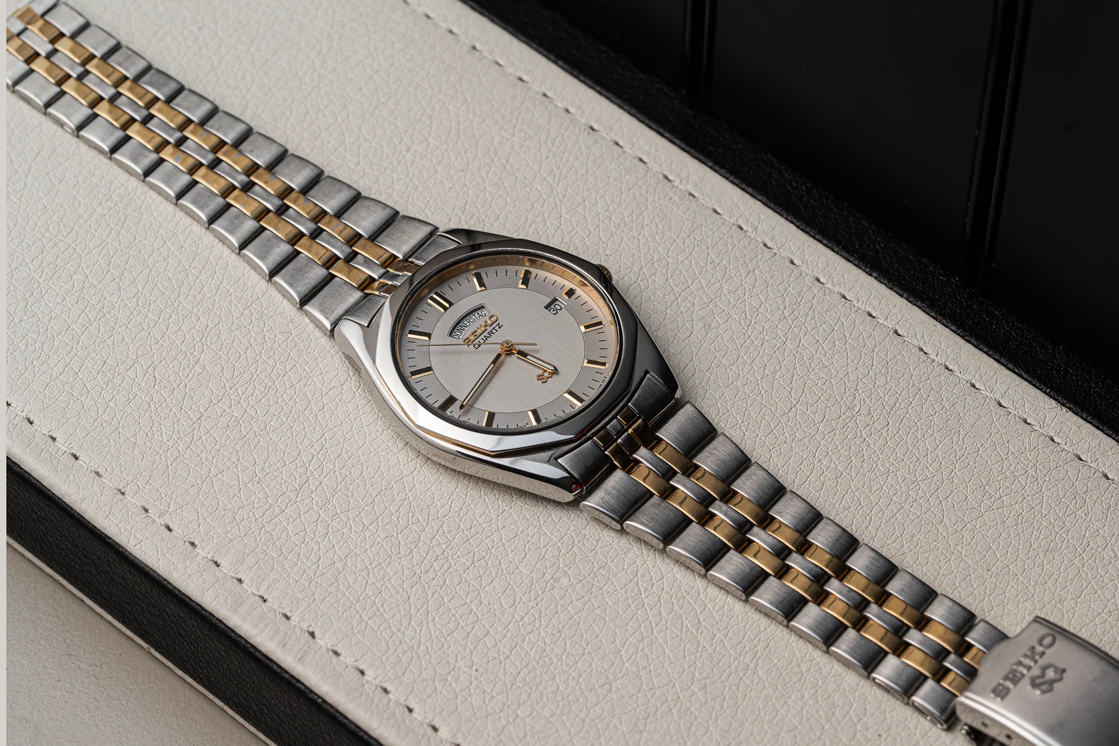 A two-tone stainless steel and gold watch Seiko SQ 5Y23-8240 with a silver dial, resting diagonally on a textured off-white leather surface with a black background.