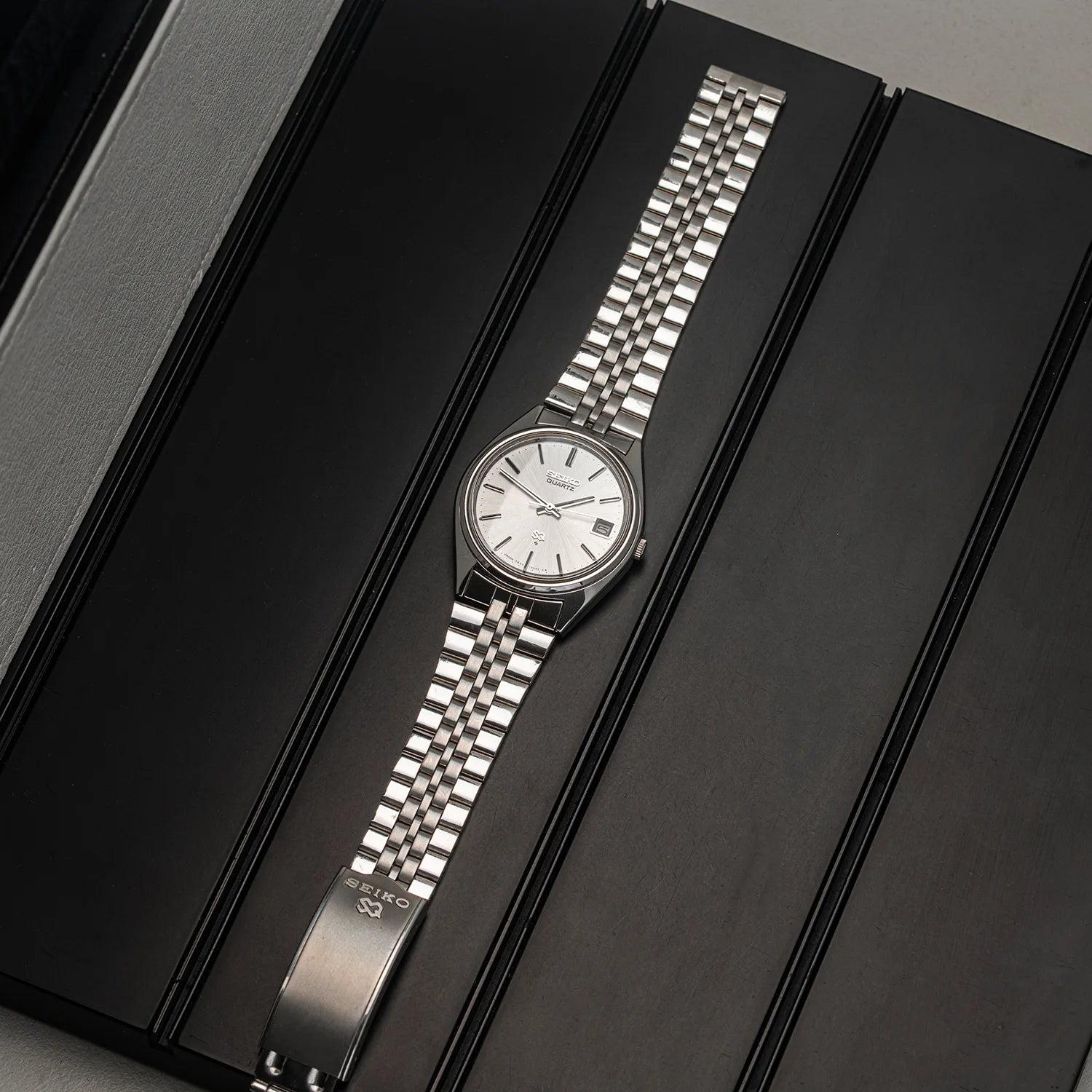 A silver-tone wristwatch Seiko SQ, Ref. 7545-7030 with a matching metal link bracelet and a white dial, displayed diagonally on a black textured surface.
