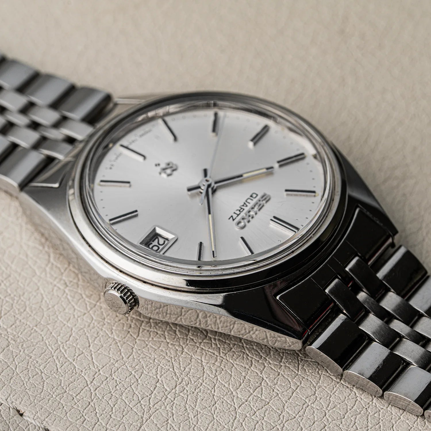 A close-up of a Seiko SQ, Ref. 7545-7030 Quartz wristwatch with a silver dial and stainless steel link bracelet, resting on a textured beige surface.