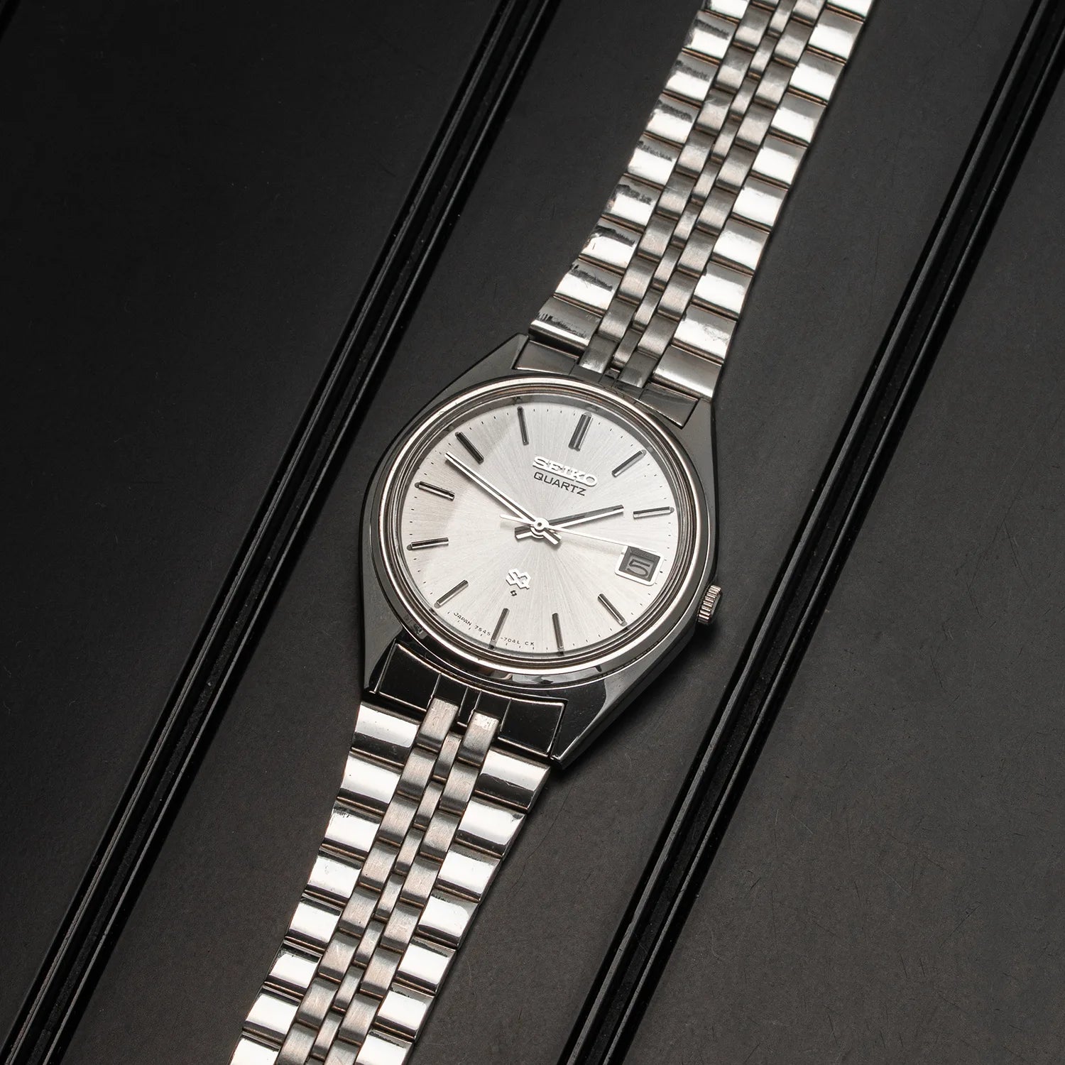 A vintage Seiko SQ, Ref. 7545-7030 watch with a silver stainless steel Oyster bracelet and a white dial, presented diagonally on a black textured background.