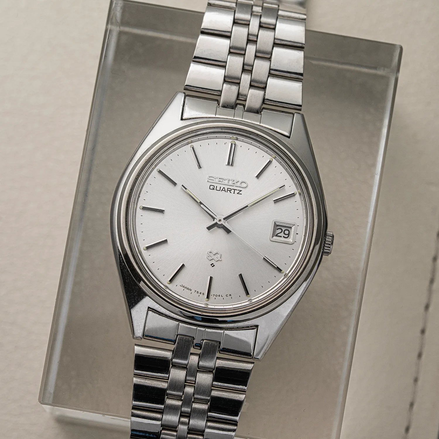 A vintage Seiko SQ, Ref. 7545-7030 Quartz wristwatch with a silver stainless steel case and link bracelet, featuring a white dial with black markings and a date window, displayed on a clear acrylic stand.