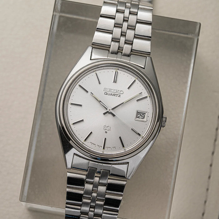 A vintage Seiko SQ, Ref. 7545-7030 Quartz wristwatch with a silver stainless steel case and link bracelet, featuring a white dial with black markings and a date window, displayed on a clear acrylic stand.