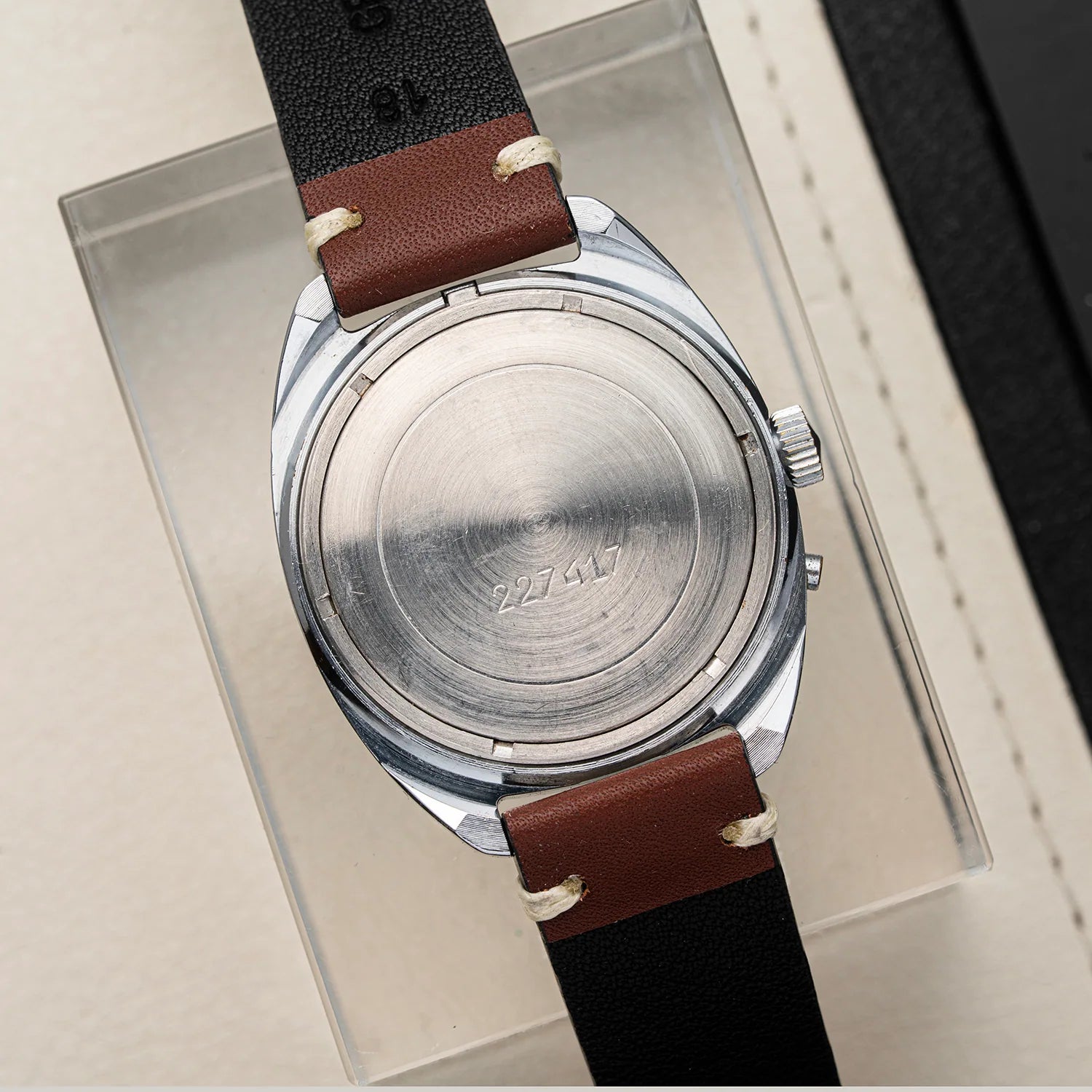 A vintage wristwatch Slava Big Numerals shown from the back, featuring a brushed silver metal case and a brown leather strap with black stitching and a black end section, displayed on a clear acrylic stand.