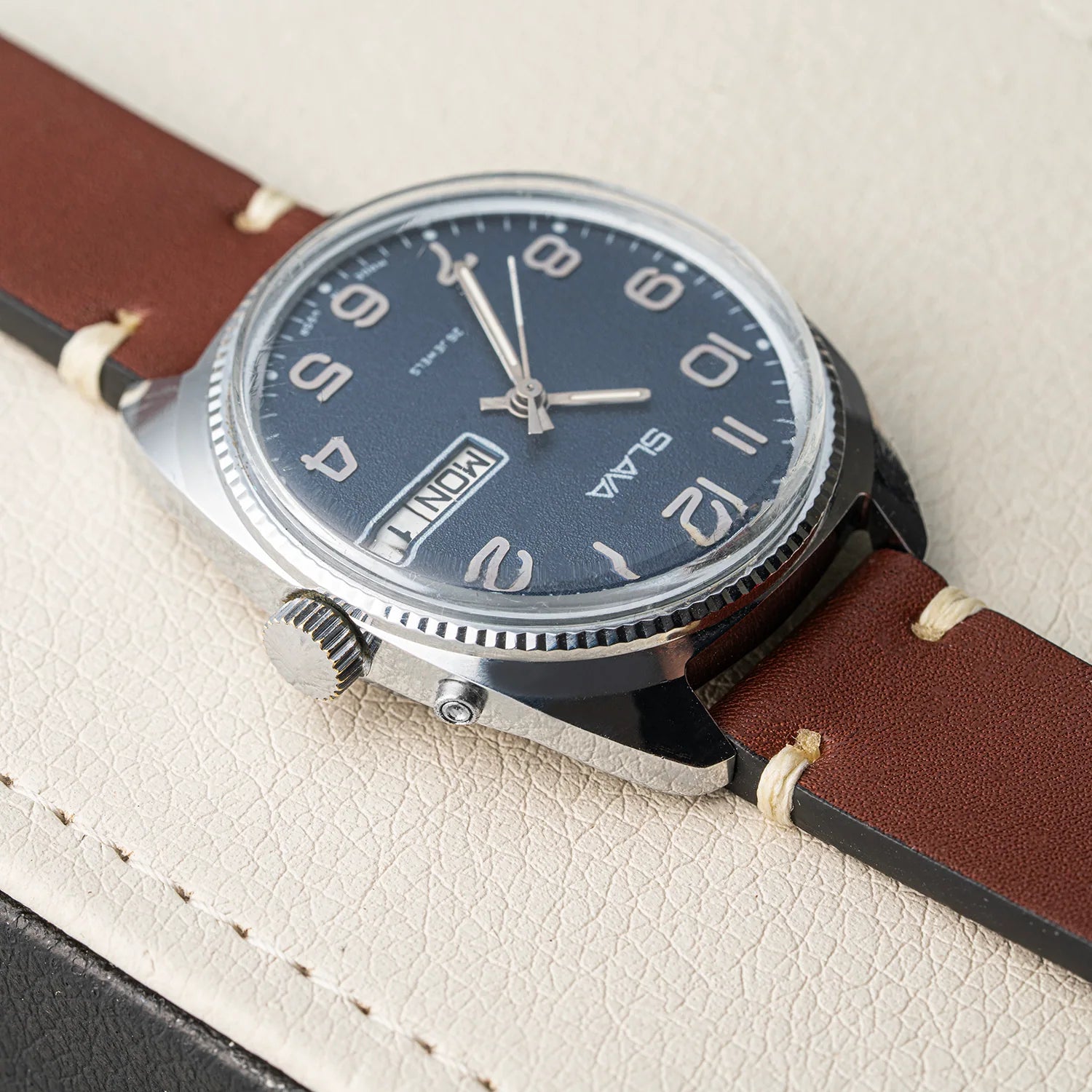 A close-up of a Slava Big Numerals wristwatch with a dark blue dial, silver-tone case, and brown leather strap, resting on a textured cream-colored surface.
