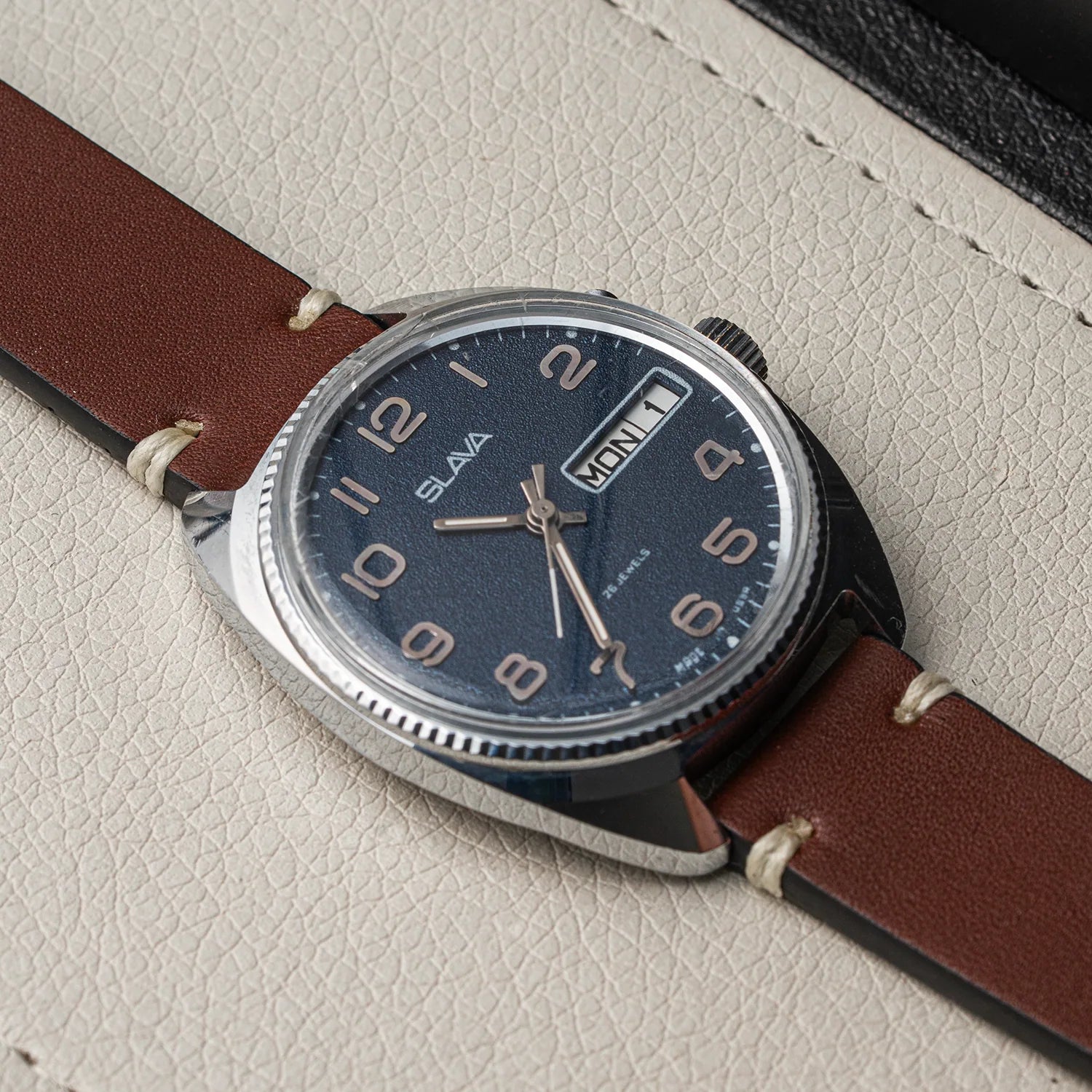 A close-up of a Slava Big Numerals wristwatch with a dark blue dial, silver-tone case, and brown leather strap, resting on a textured light beige surface.