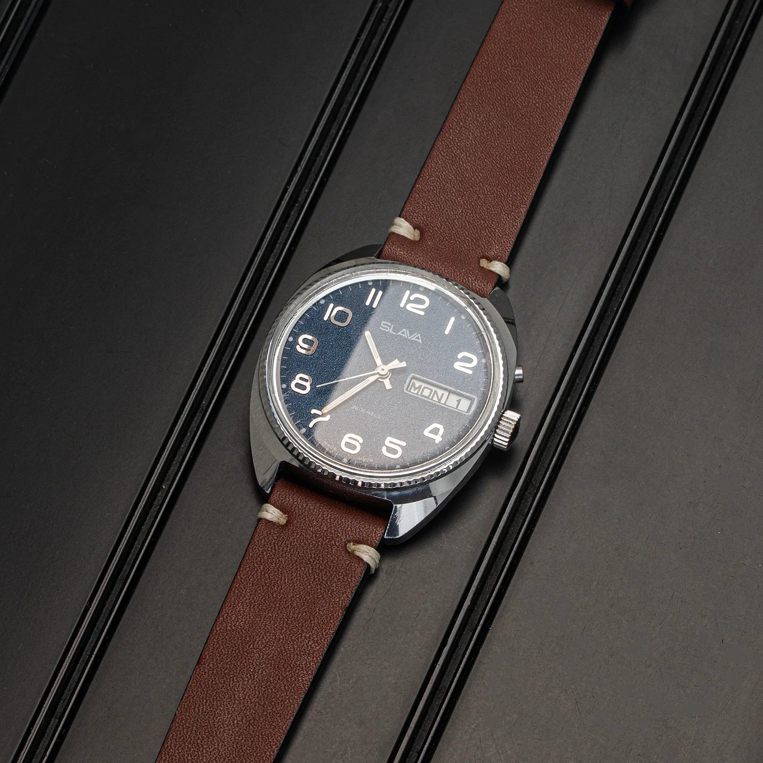 A wristwatch Slava Big Numerals with a brown leather strap, a silver-toned metal case, and a dark blue dial with white Arabic numerals, displayed diagonally on a dark textured background.