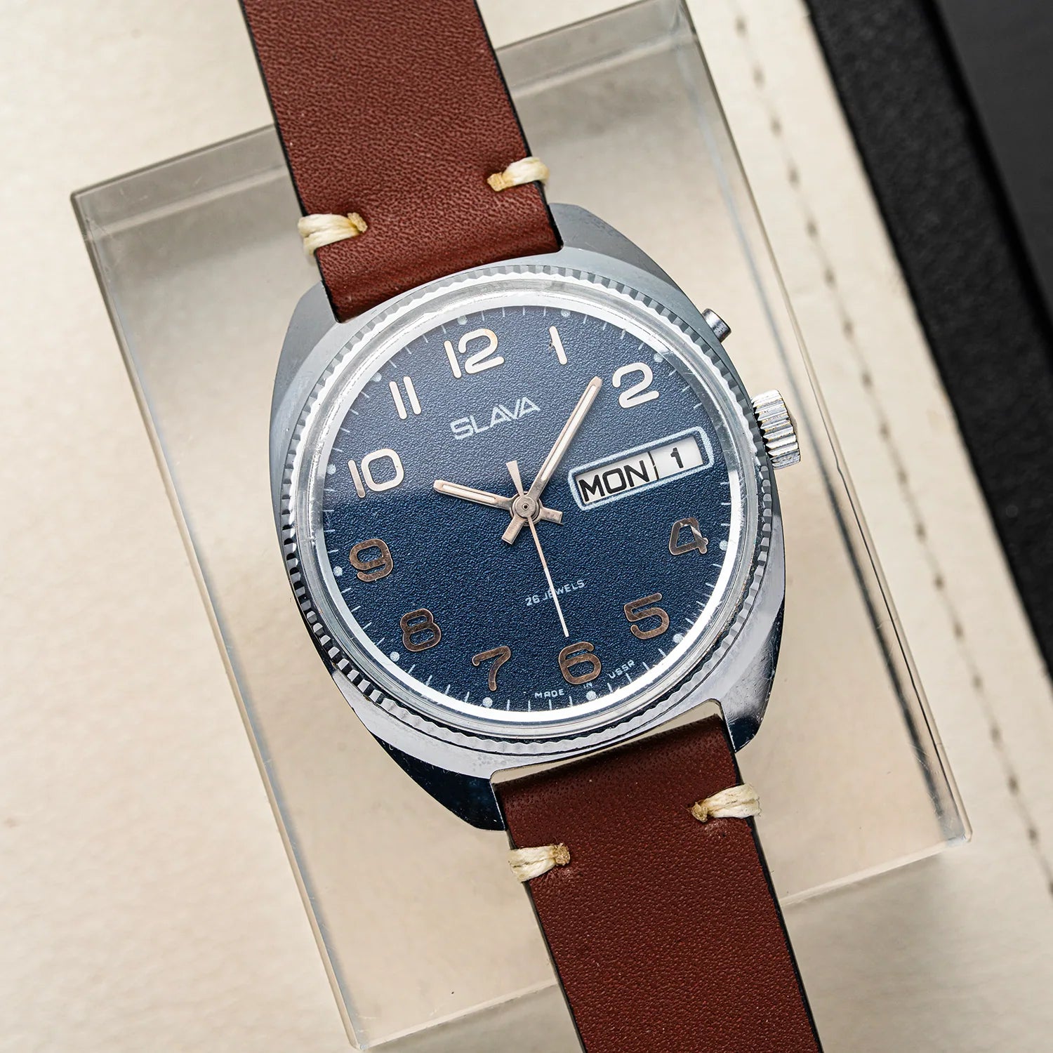 A vintage-style wristwatch Slava Big Numerals with a brown leather strap, a silver-toned case, and a dark blue dial. The watch is displayed inside a clear acrylic box on a beige surface.
