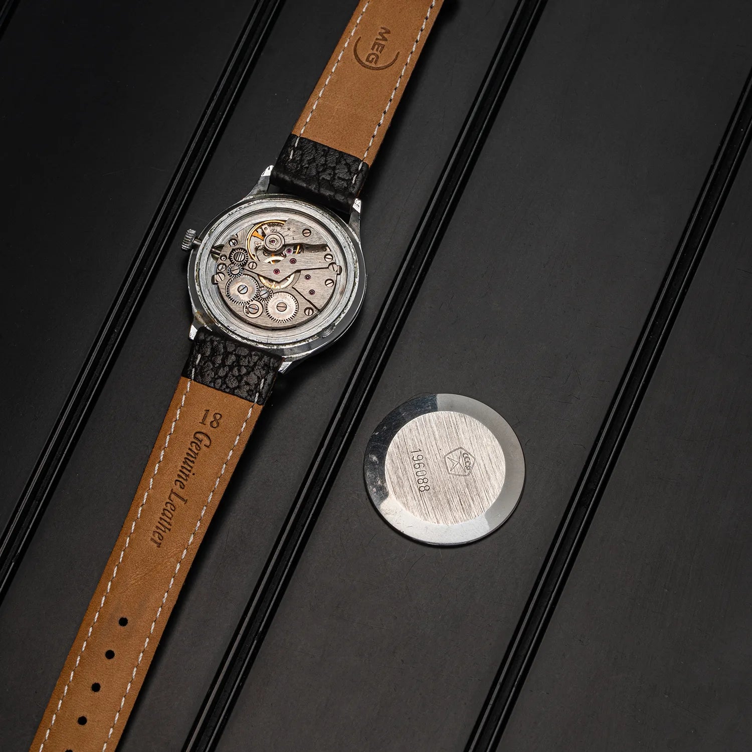 A luxury watch with a brown leather strap and a silver-toned movement visible through the open case, displayed diagonally on a dark textured background next to a round silver metal emblem.
