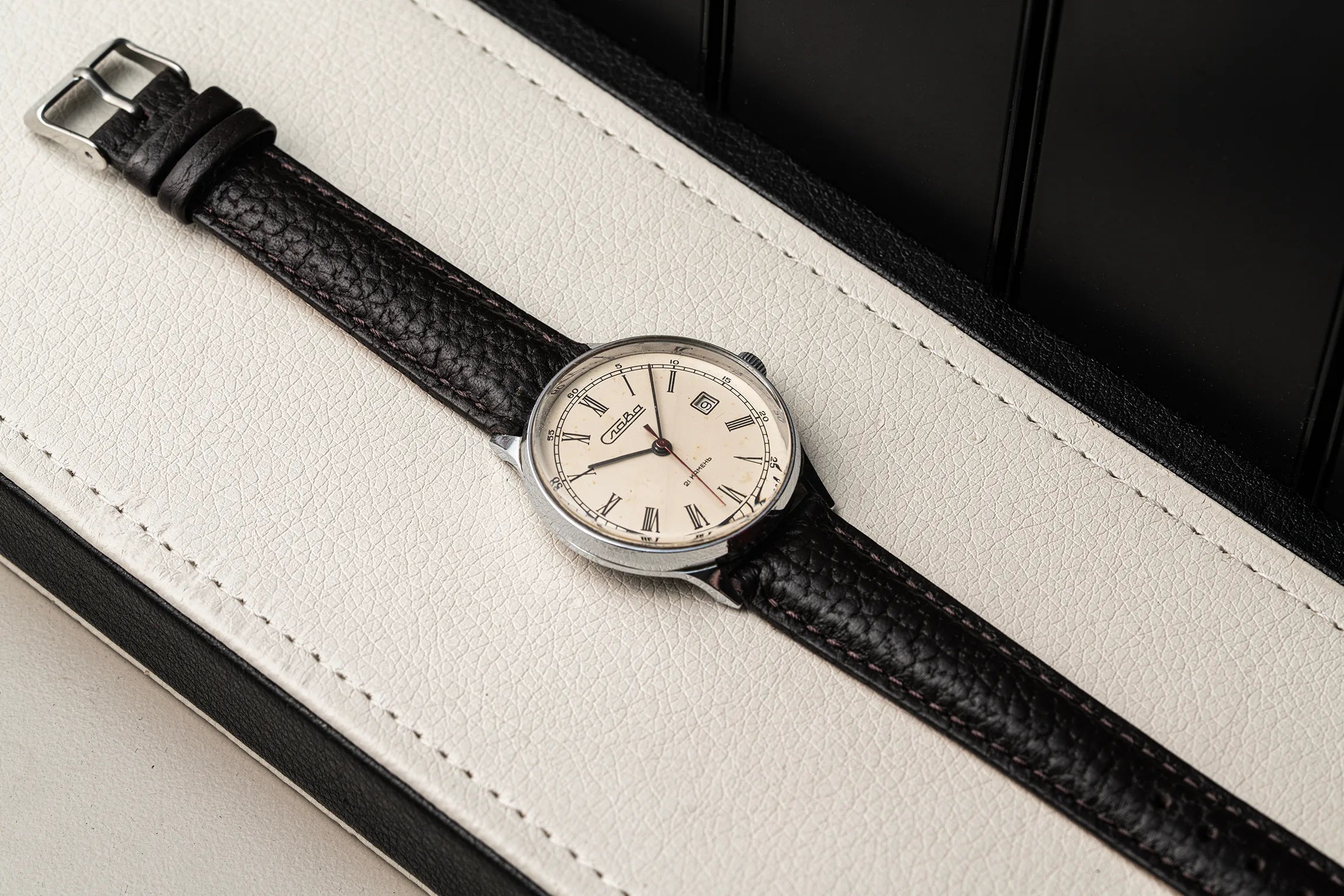 A wristwatch Slava Roman Numerals with a black textured leather strap and a silver-toned round dial, resting diagonally on a white leather surface with a black background.