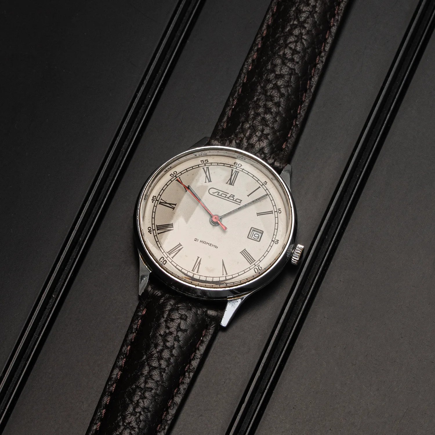 A vintage-style wristwatch Slava Roman Numerals with a silver-toned oval case, cream-colored dial, and a textured dark brown leather strap, displayed diagonally on a dark textured background.